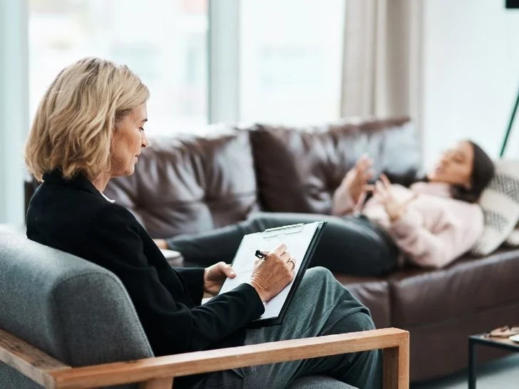 therapist in chair taking notes with client on sofa talking therapy room session