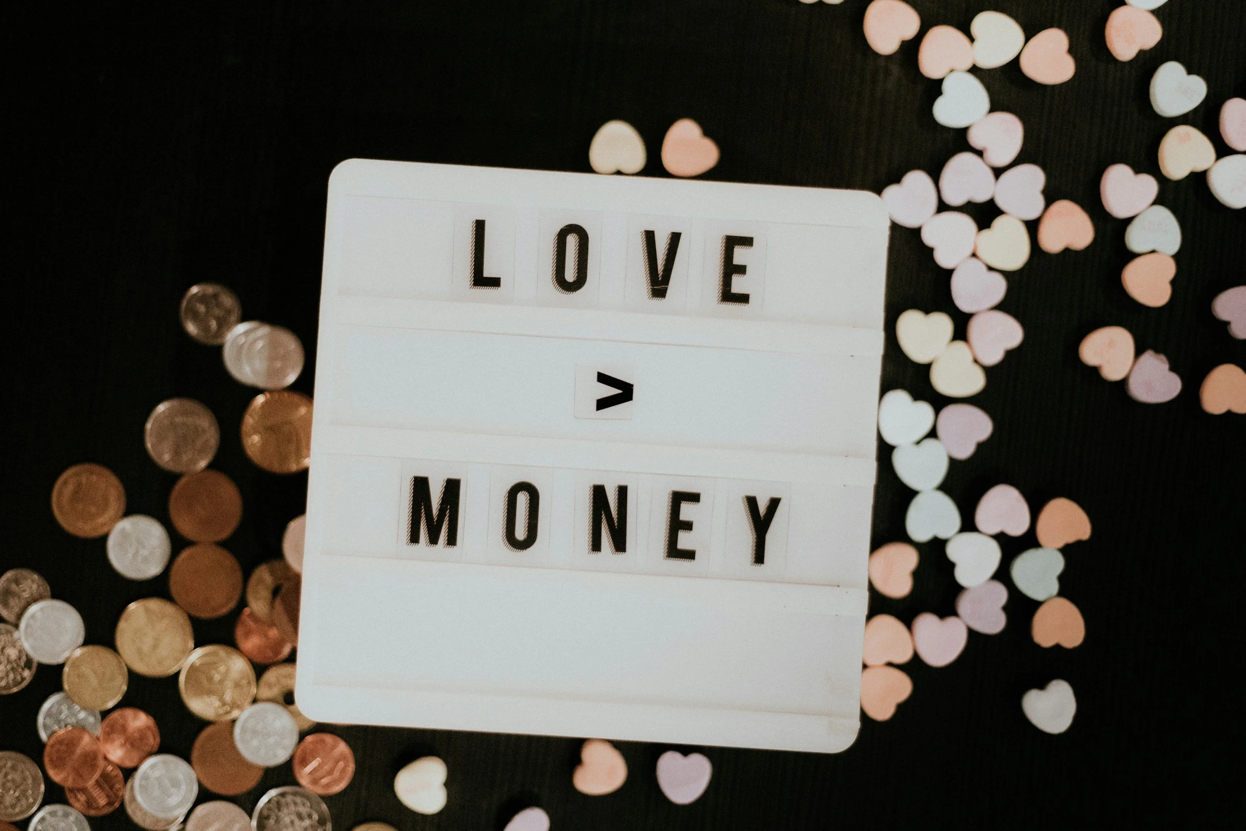 sign that says love > money with different colored coins scatteres across black background