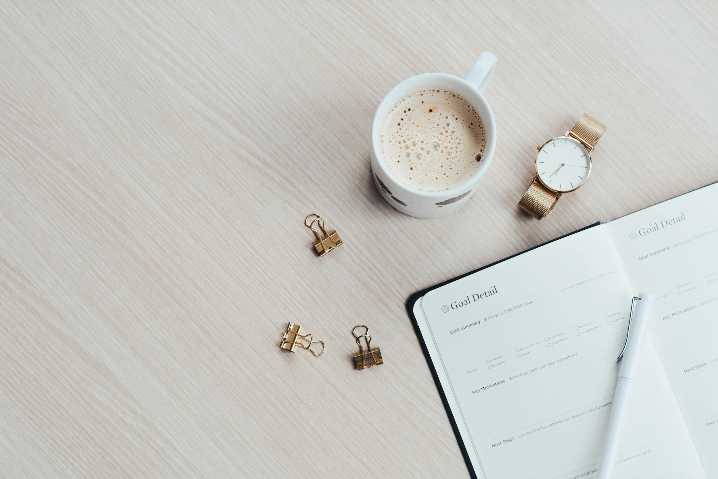 Goal jounral open with white pen laid out with a mug of hot chocoloate and a gold watch anda 3 gold pin clips surrounding it