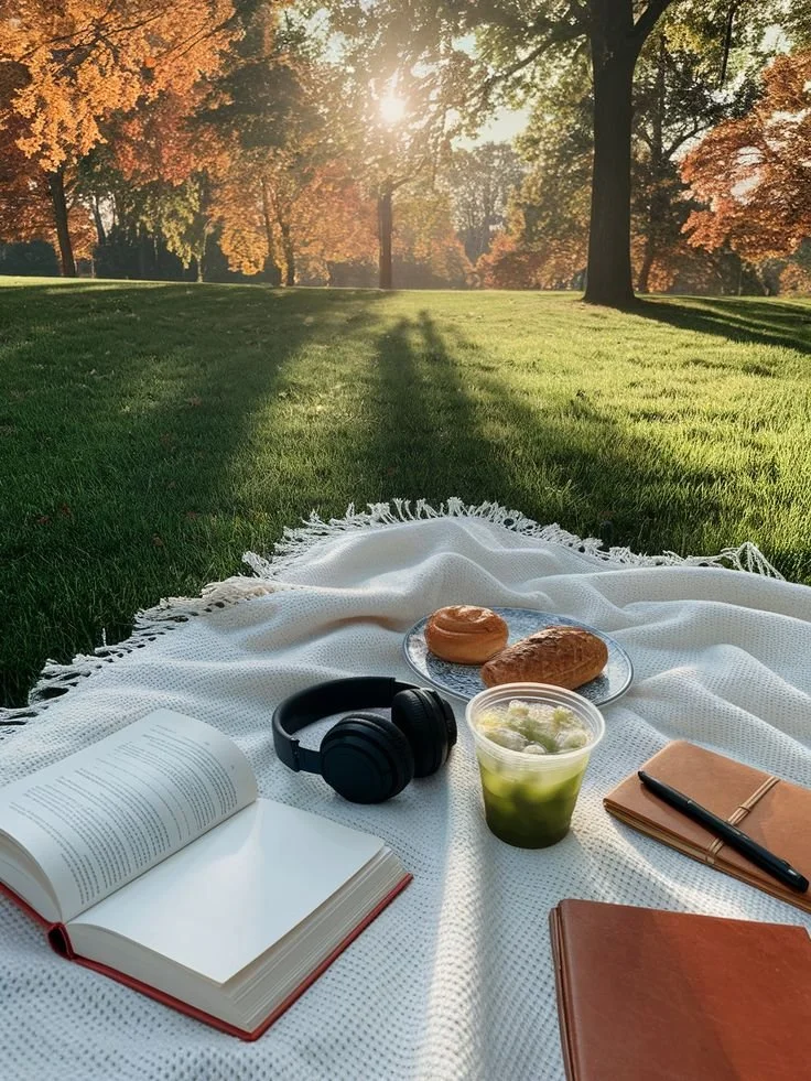 outdoor image in a park in the fall with view of white picnic blanket and plate of breads, headphones, iced matcha, journal with pen and open book with sunlight peeking through centre field
