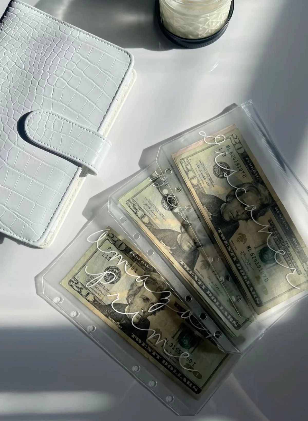 3 clear enveloped with category written ontop in cursive with twenty dollar bills sitting inside next to a white crocodile textured wallet