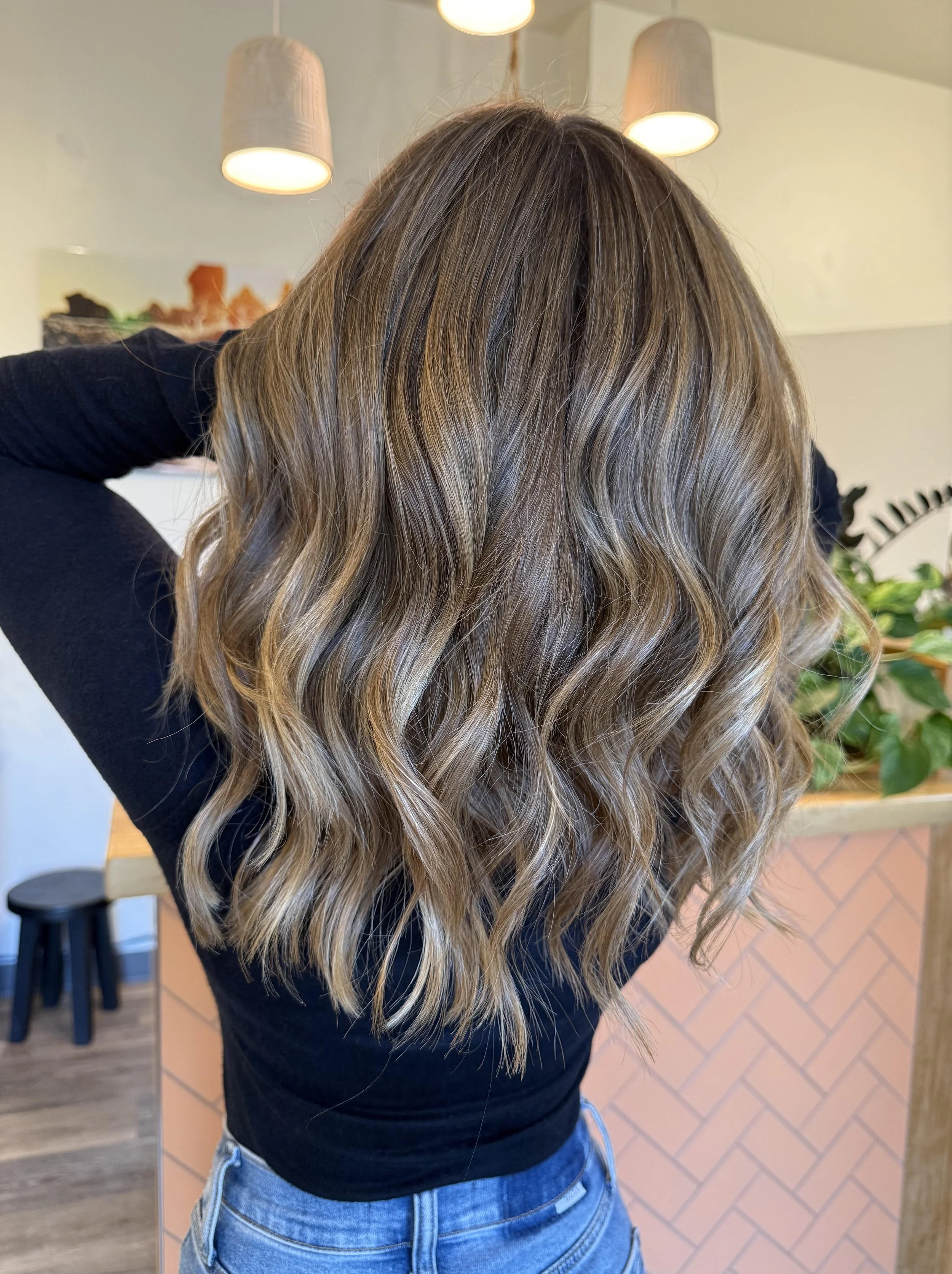 Woman with wavy, highlighted hair in a salon or café setting, facing away from the camera.