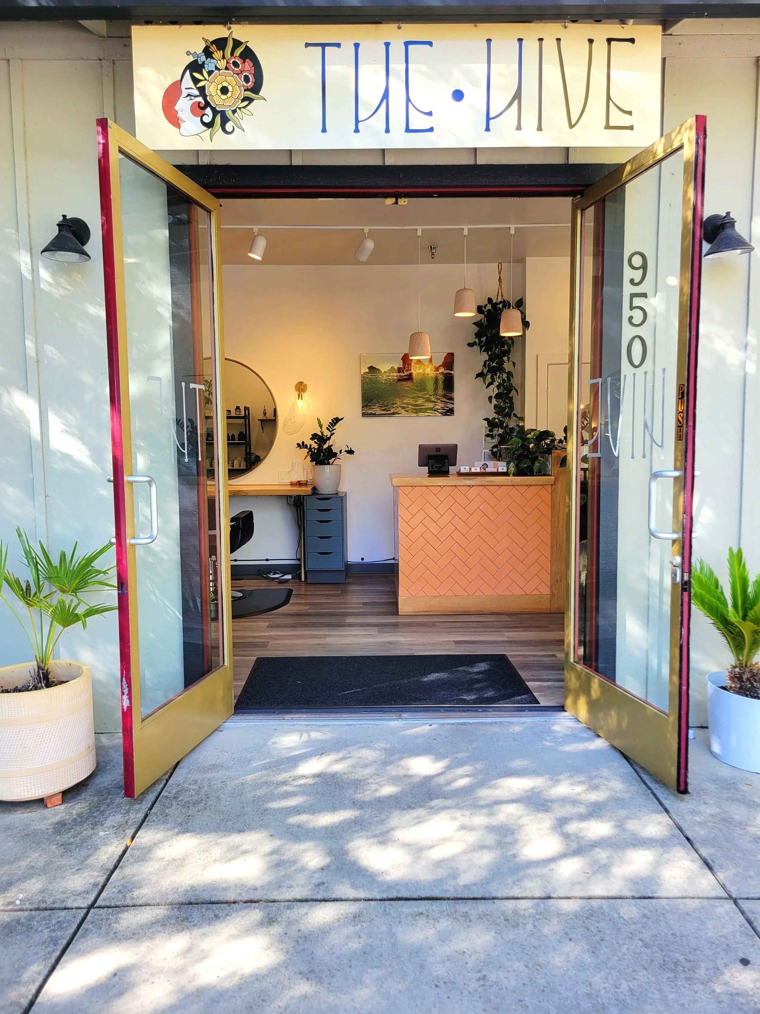 Entrance to a modern salon named 'The Hive,' with open glass doors, interior decor including a reception desk, plants, lighting, and wall art. The address '950' is visible on the right side of the door.