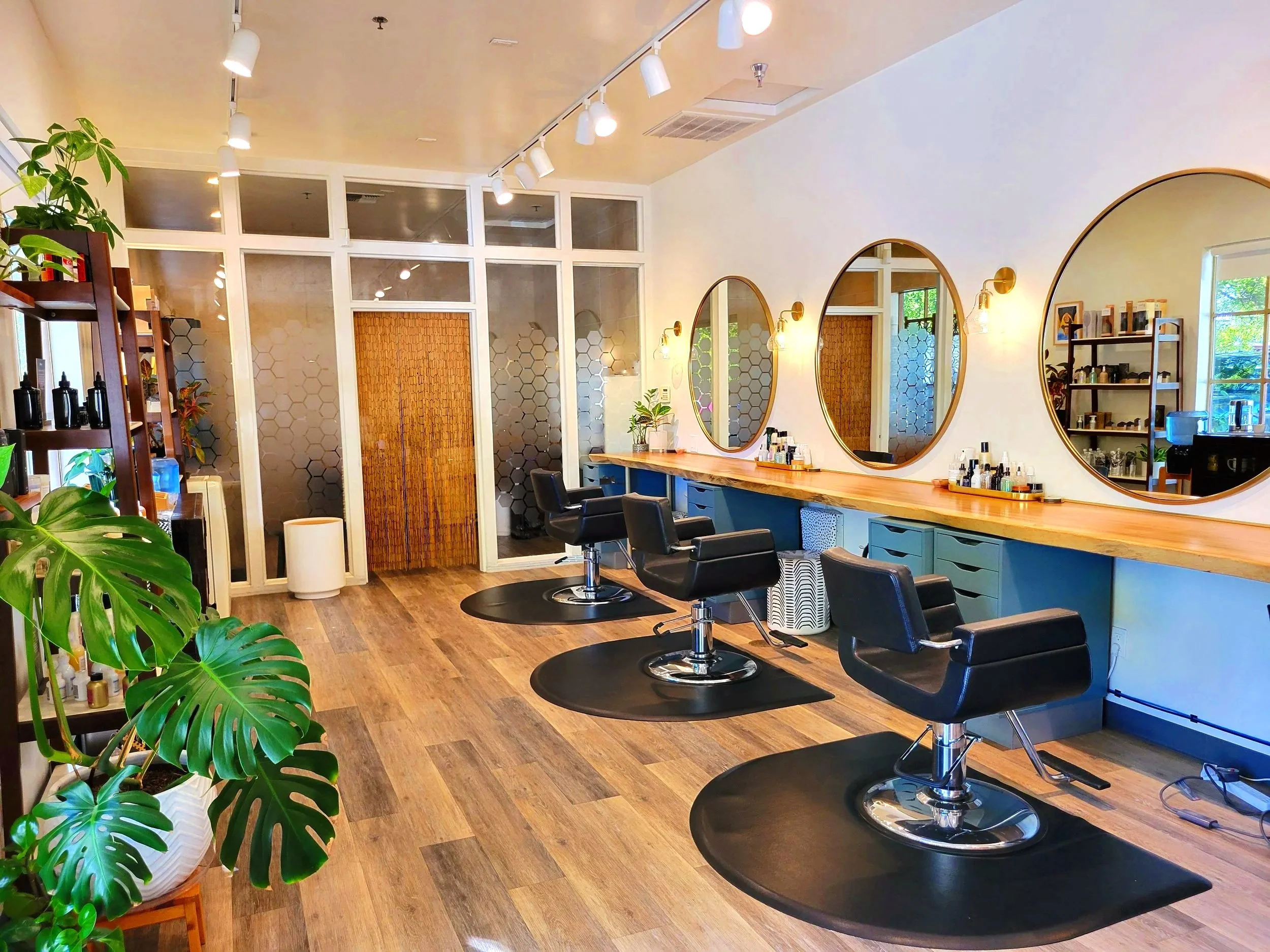 Modern hair salon with three black styling chairs, large round mirrors, wooden countertops, and shelves with hair products. The salon has wooden flooring, plants, and large windows allowing natural light.