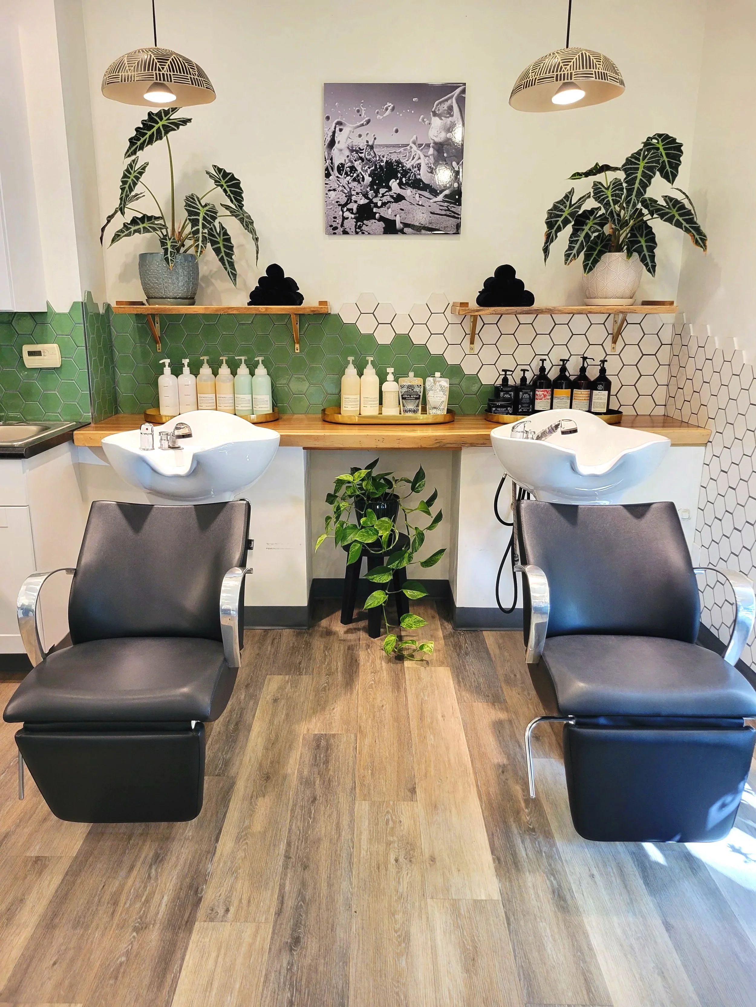 A modern salon station with two black chairs in front of a wooden counter, two white sinks, and shelves holding plants and toiletries, with a wall-mounted black-and-white art piece and decorative lighting.
