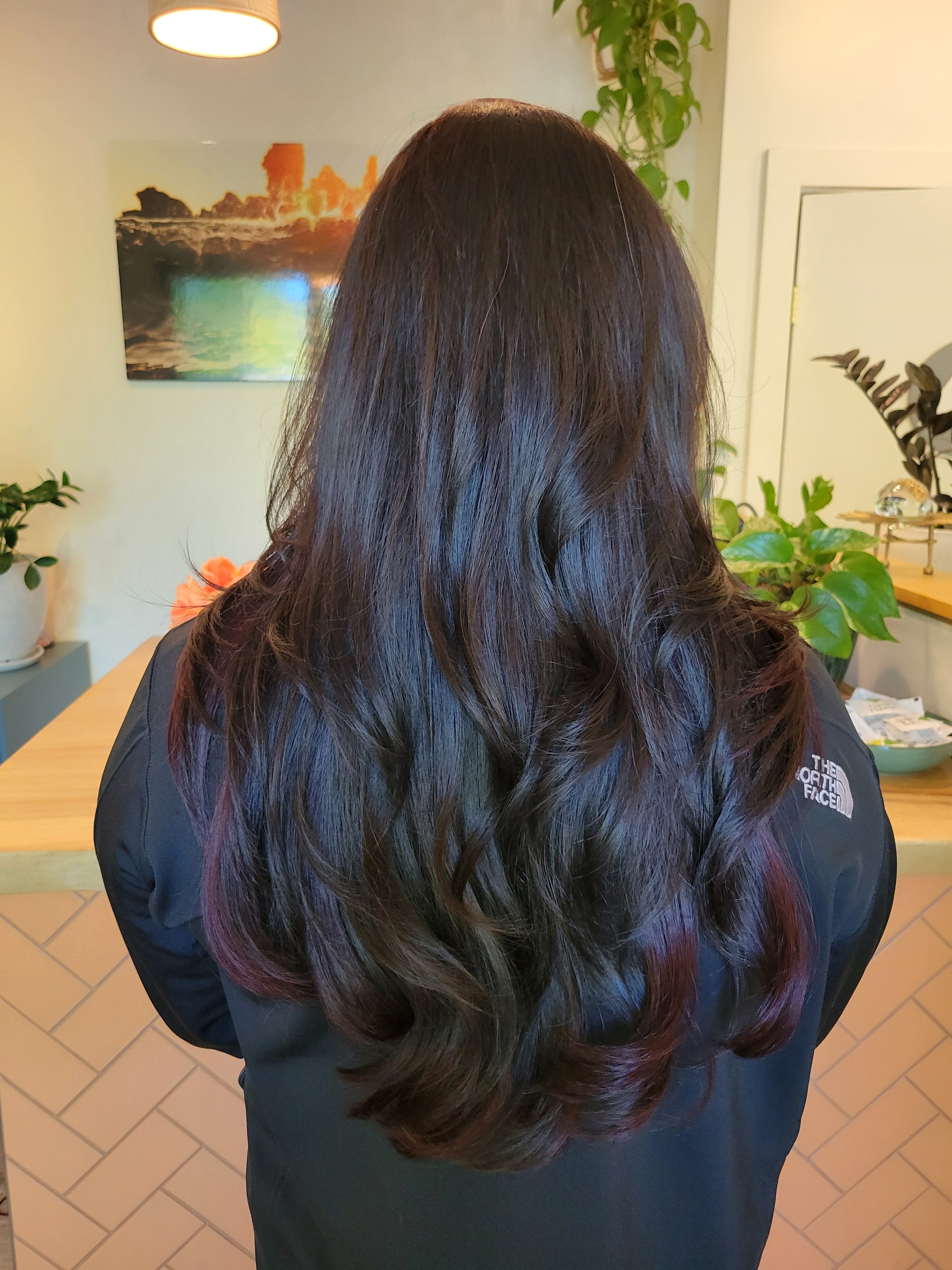 A woman with long, wavy dark hair with red tips, wearing a black North Face jacket, standing indoors with her back to the camera, in a room decorated with plants and wall art.