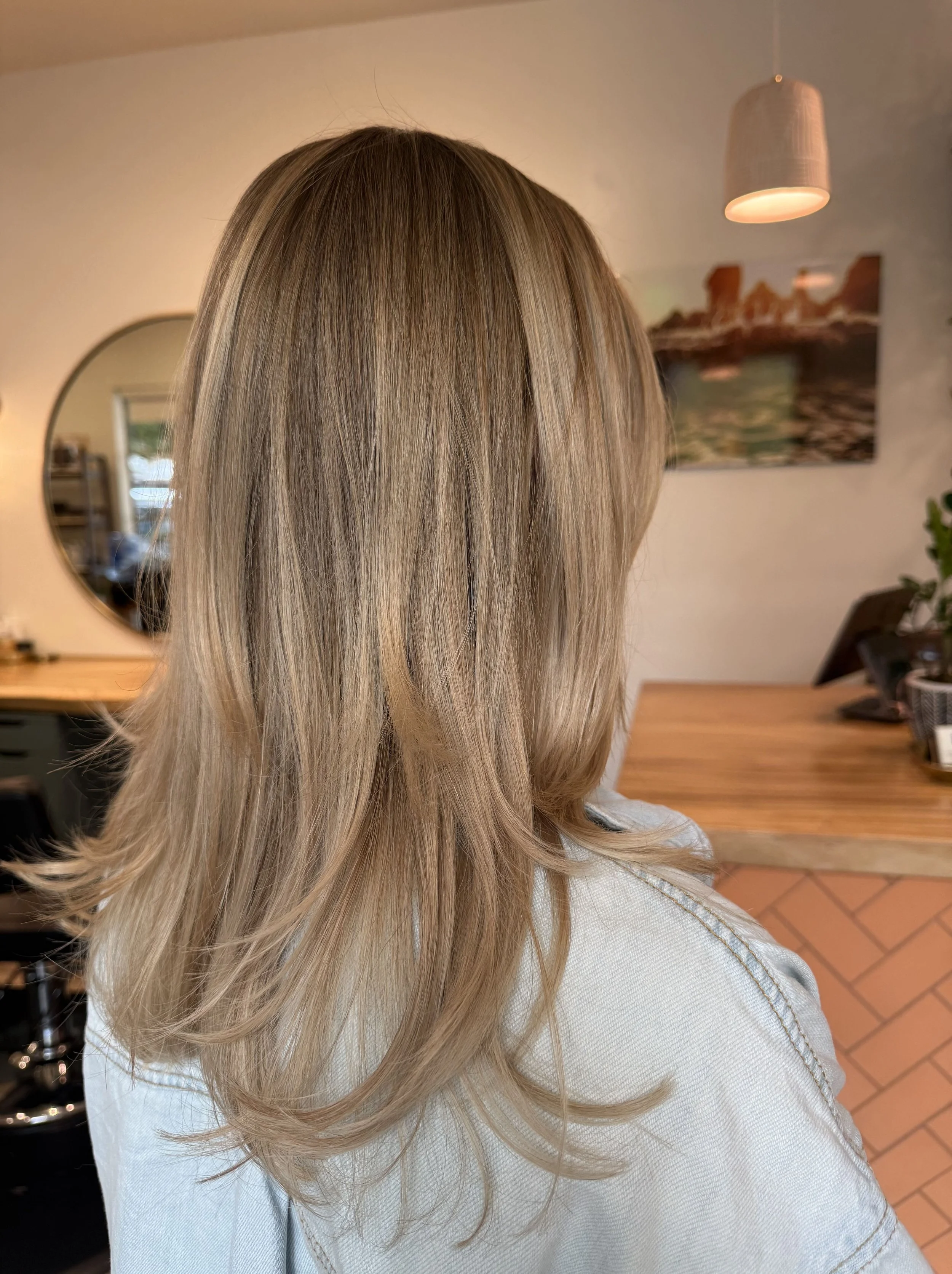 Back view of a woman with shoulder-length, blonde hair styled in loose layers in a hair salon.