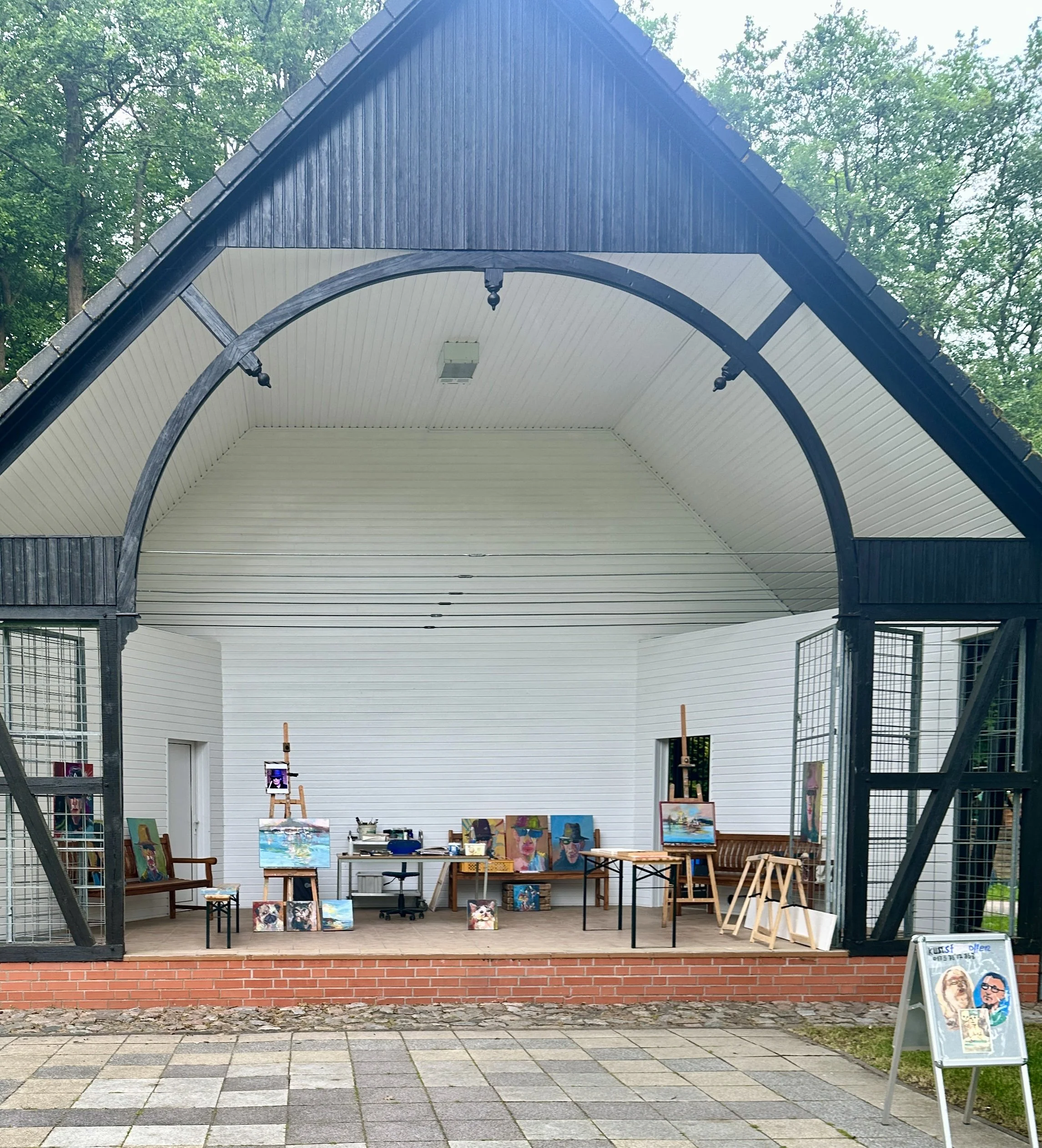 Eine offene Galerie im Freien mit Malereien auf Leinwand, die auf Ständern und auf dem Boden ausgestellt sind. Es gibt Sitzgelegenheiten, einen Galerie-Host-Stand und eine Whiteboard-Tafel. Die Galerie ist im Schatten eines Daches auf einer Steinterrasse mit Ziegelmauer gebaut, umgeben von Bäumen.