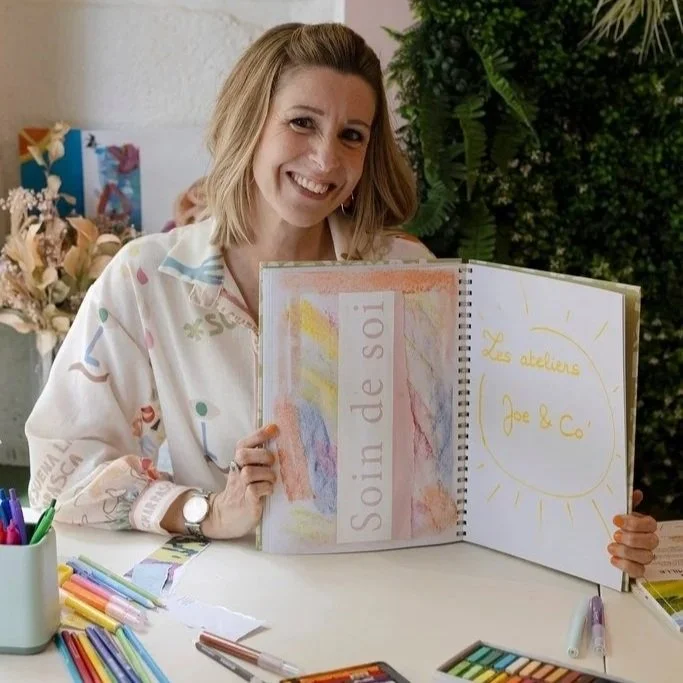 Une femme souriante tient un cahier ouvert avec des dessins et des textes en français, devant un fond de plantes vertes.
