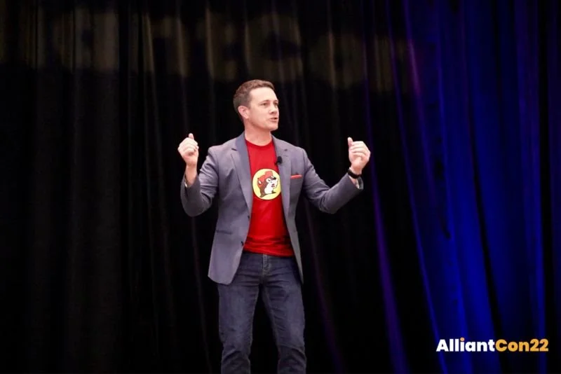 Jeff Toister on stage, making a triumphant gesture. He is wearing a red Buc-ee's t-shirt under a blue blazer.