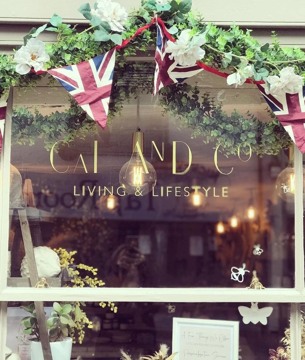 Shop window decorated with greenery, white flowers, and Union Jack flags, featuring the sign 'Cai & Co Living & Lifestyle'.