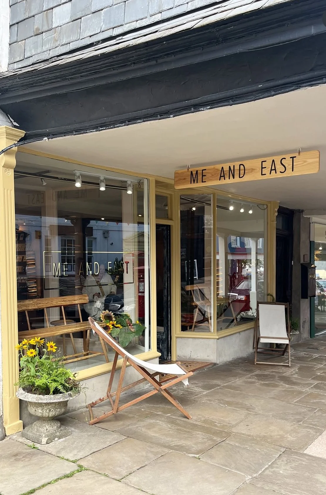 Storefront with a wooden sign that says 'ME AND EAST' above the entrance, large windows displaying the shop's name, outdoor chairs, and potted flowers.