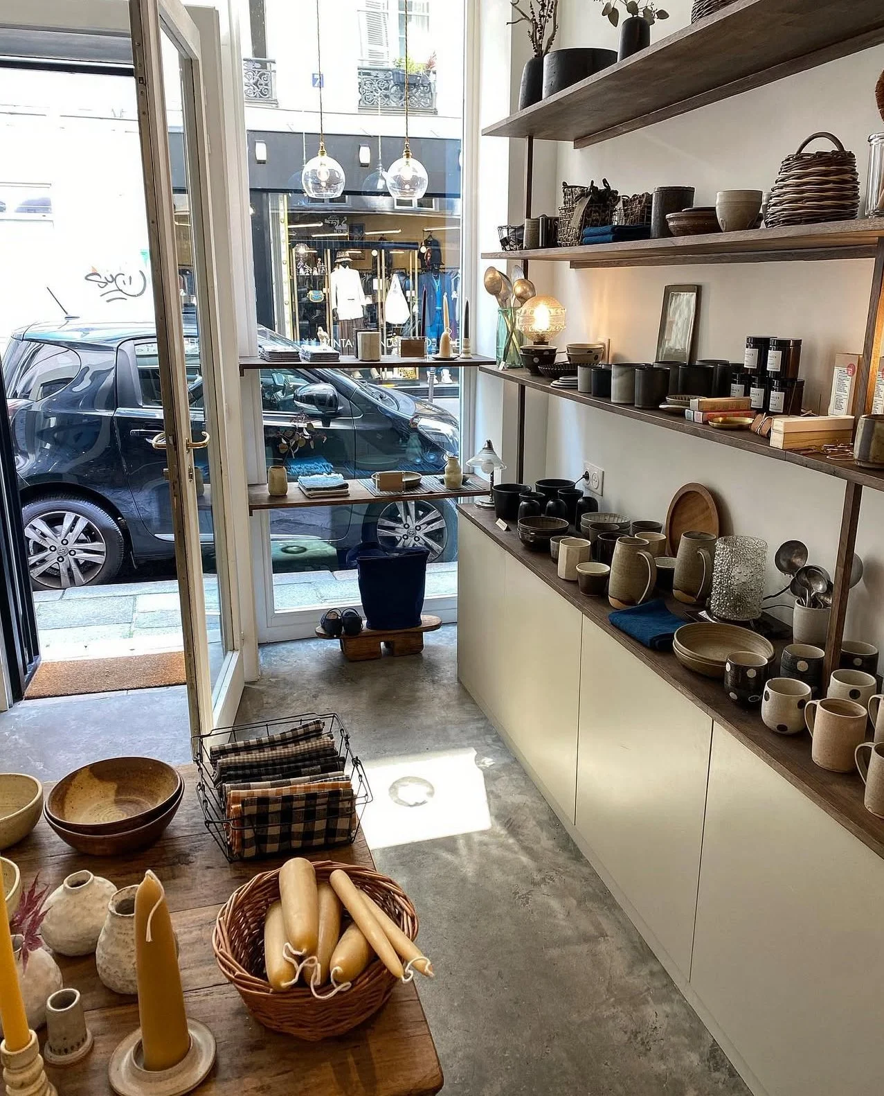 Interior of a boutique store with shelves displaying ceramic and pottery items, plates, cups, bowls, and decor. Wooden shelves line the white wall, and a wooden table in the foreground has candles, bowls, and a basket of candles. Large glass door and window reveal a street scene with parked cars and mannequins outside.