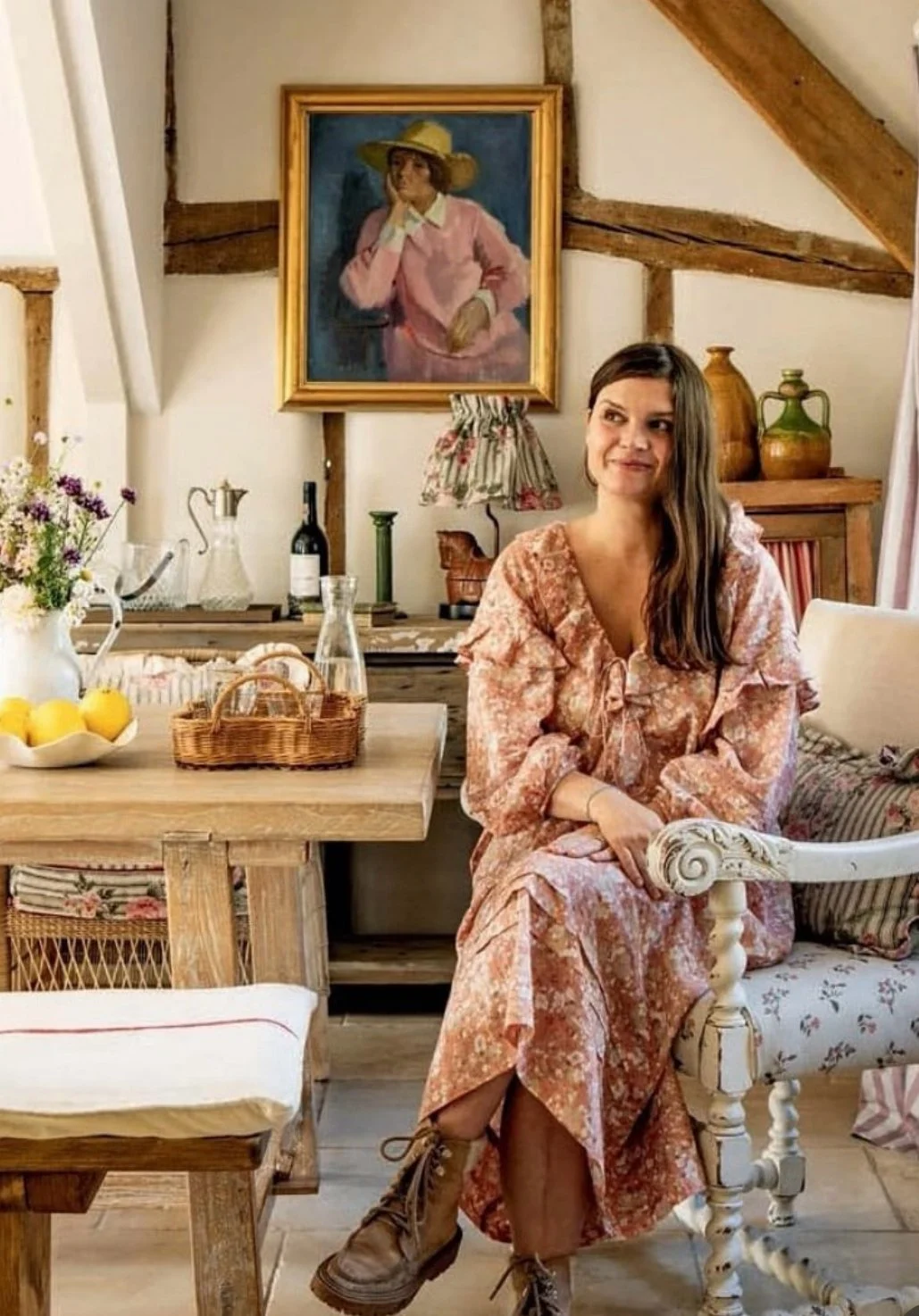 A woman with long brown hair in a loose braid, wearing a pink floral dress and brown lace-up boots, sits on a vintage white and floral armchair in a cozy, rustic room. Behind her is a wooden sideboard with a lamp, vases, and bottles, above which hangs a painting of a woman in a pink shirt and wide-brimmed hat. The room features wood beams and a wooden table with a basket of lemons, a wine bottle, and a glass decanter.