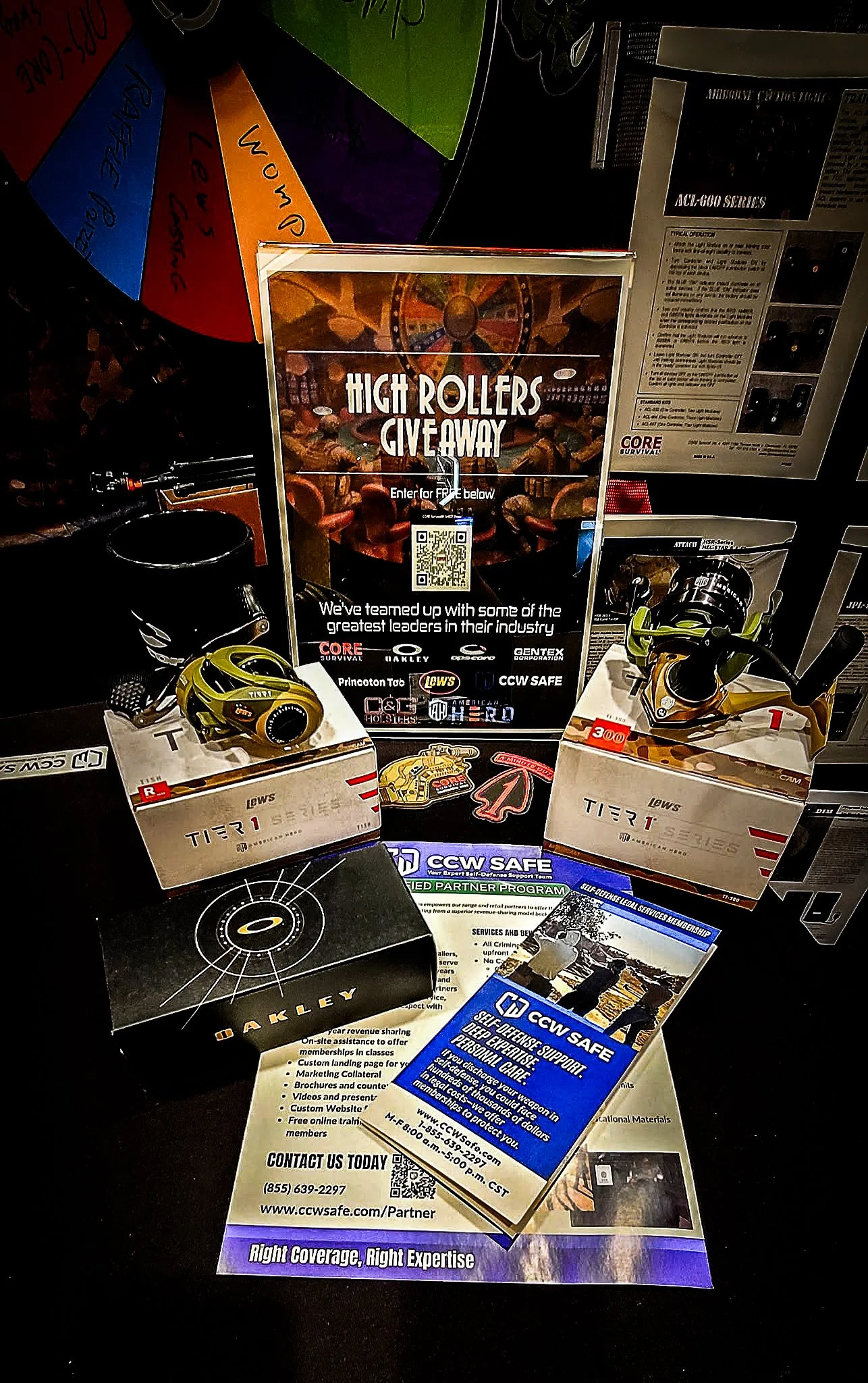 Table display with promotional materials for a high rollers giveaway event, featuring small boxed items, Flyers, and signage from brands like Oakley and Leupold, along with informational cards about CCW support services.