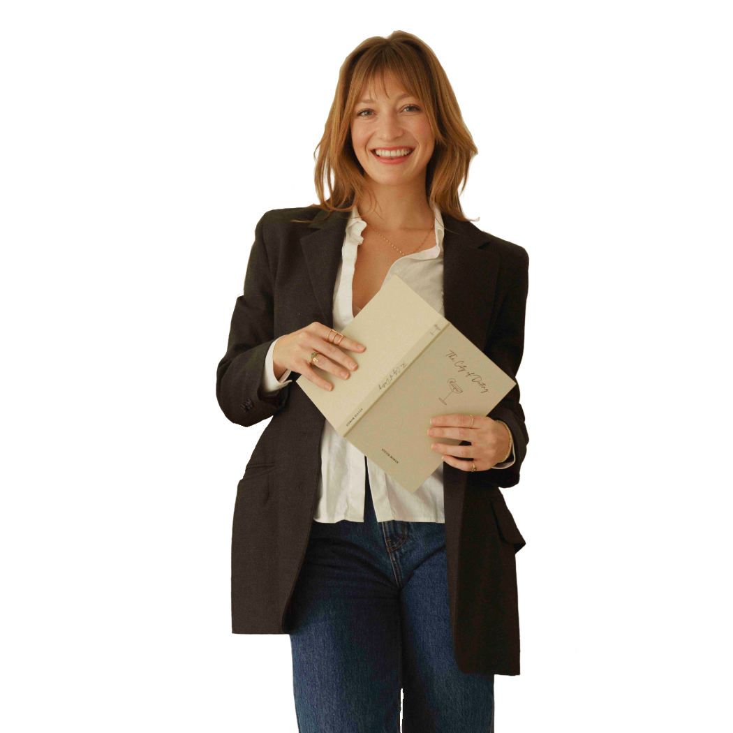 A woman with shoulder-length red hair wearing a black blazer and white shirt, smiling and holding an open book titled 'The Art of Poetry' against a plain background.