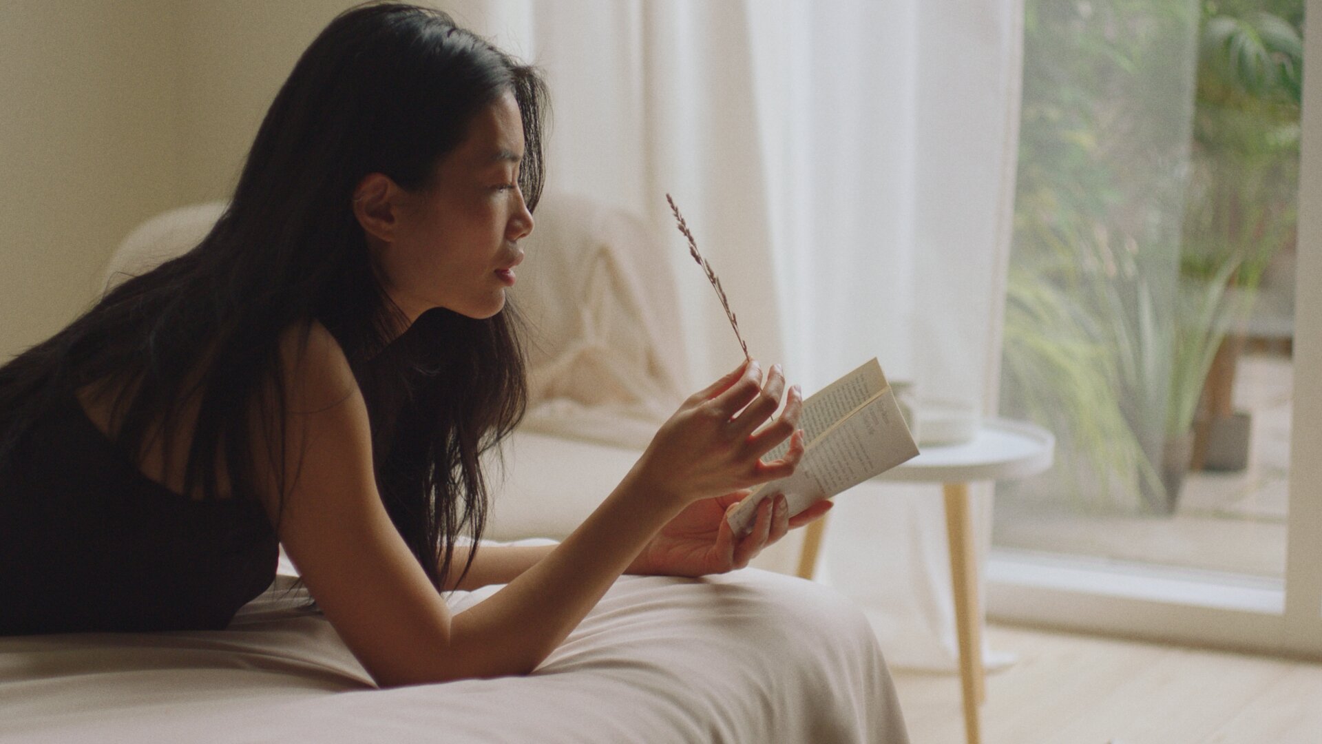 A woman lays on a bed outstretched reading a book and holding a flower, The Kiln video production