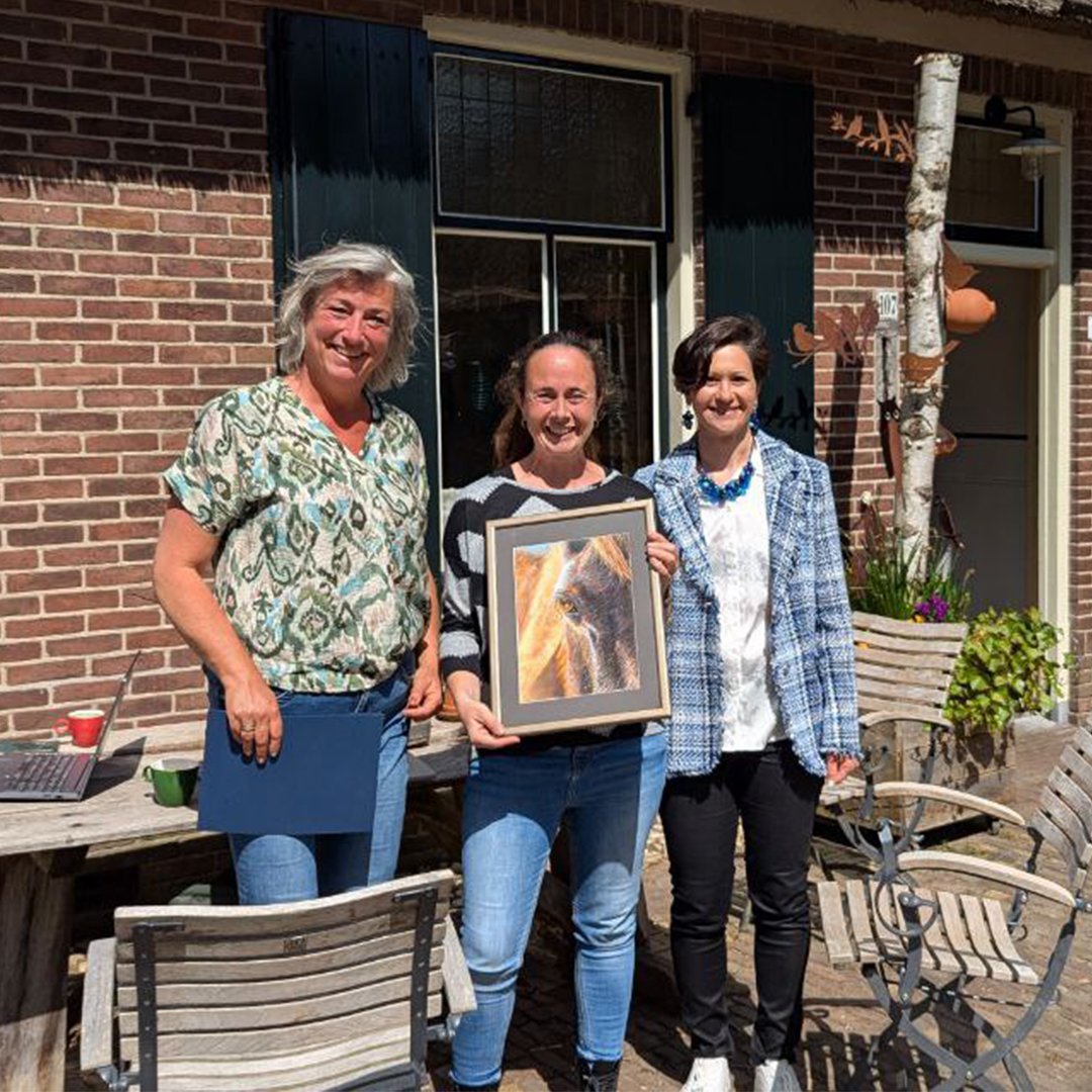 Three women standing outside on a patio, one holding a framed painting of a horse. They are smiling and appear to be celebrating. The background shows a brick wall, windows with dark shutters, and outdoor patio furniture.