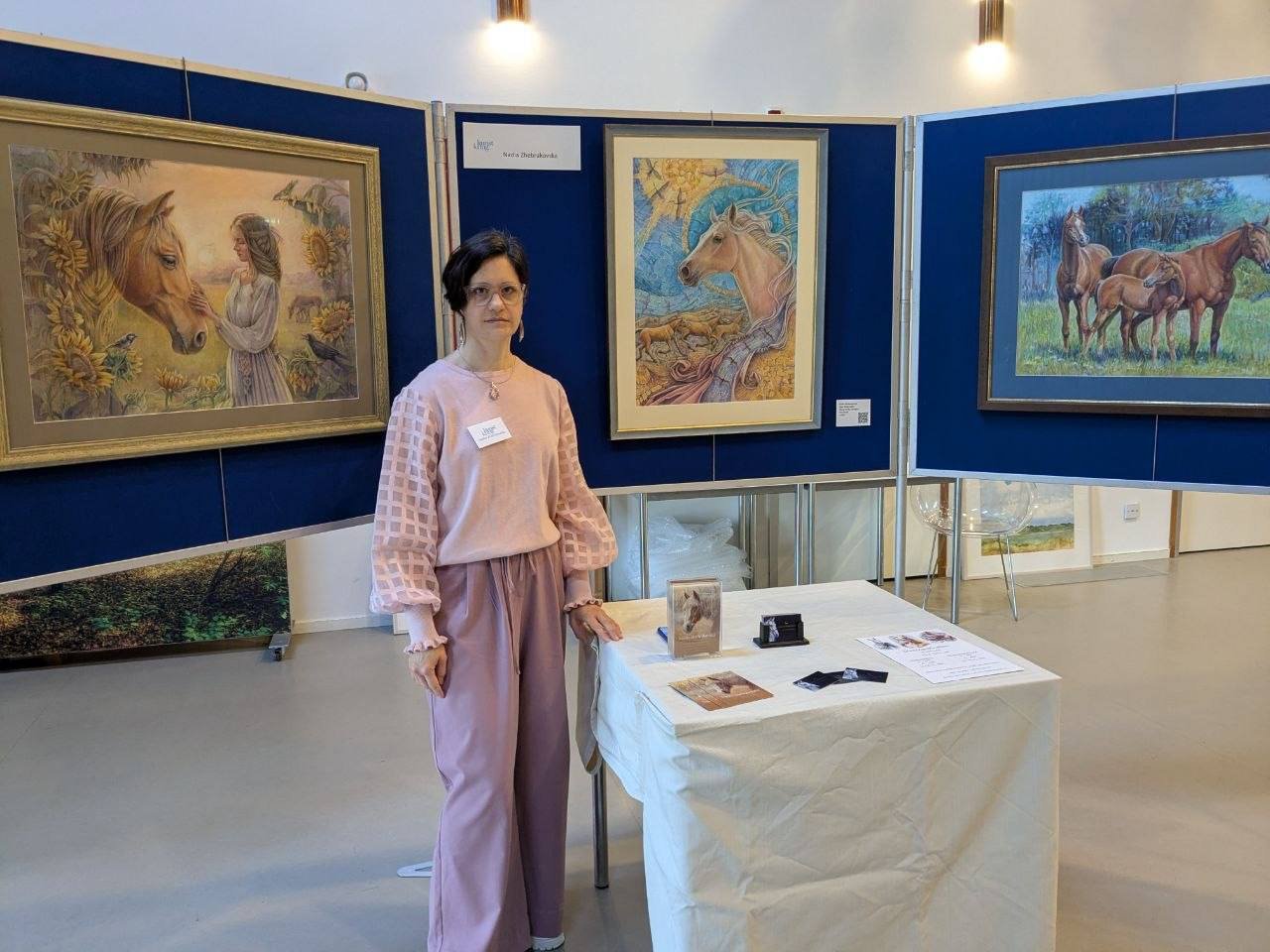 A woman stands at an art exhibit featuring three paintings of horses. She is wearing pink pants and a pink top. The paintings depict horses in various settings, with the middle painting showing a horse with flowing hair near water. There is a white t