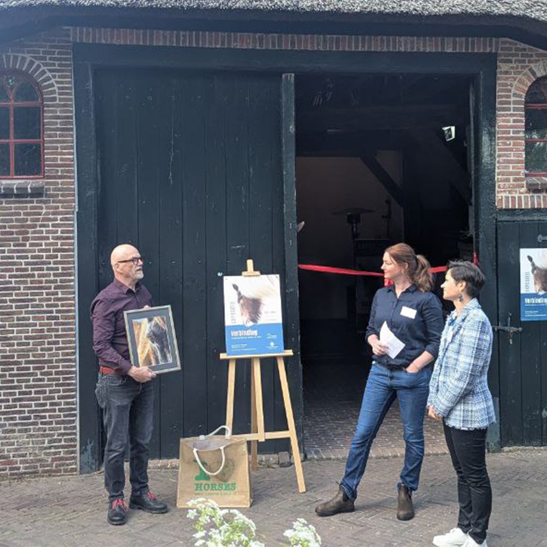 Three people standing outside a barn entrance, with one person holding a framed picture of a llama, and a poster on an easel advertising an event involving llamas, during daytime.