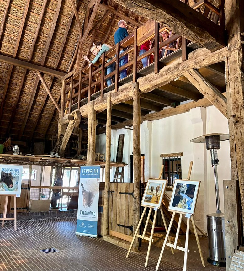 Interior of a rustic barn with wooden beams and a loft. Several people are standing on the upper level, looking down. Artistic photographs are displayed on easels and a banner stands near the center.