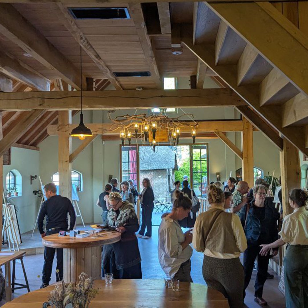 People gathering in a spacious, rustic-style event space with wooden beams, chandelier, and natural light from large open doors, attending an art exhibition.