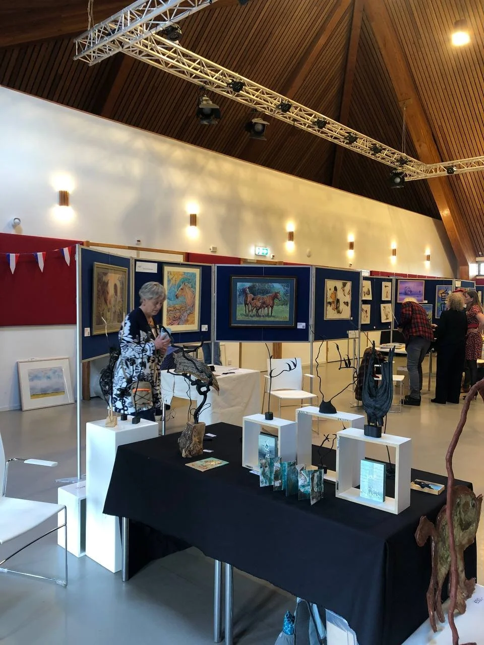An art gallery exhibition with paintings of horses and wildlife on display, people viewing the artwork, and sculptures on tables in a spacious hall with wooden vaulted ceilings.