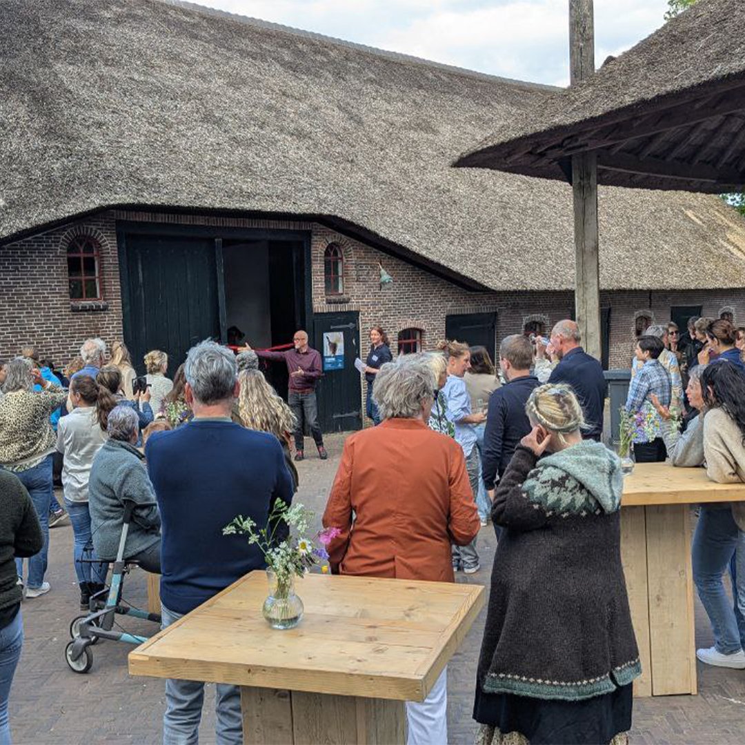 A crowd gathered outside a building with a thatched roof for an event, with some people talking and others listening as a person cuts a ribbon.