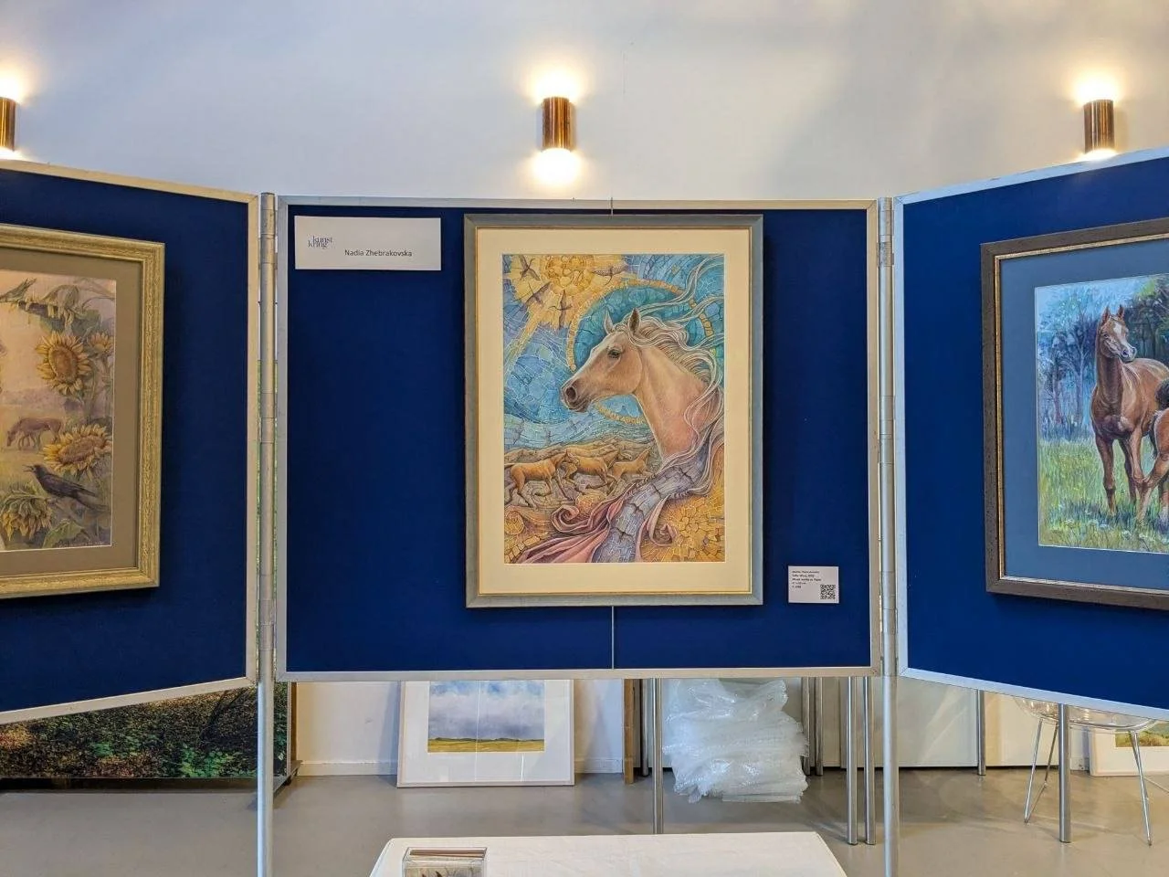 Art exhibition with paintings of horses on display in a gallery setting, blue panels, framed artwork, and overhead lights.
