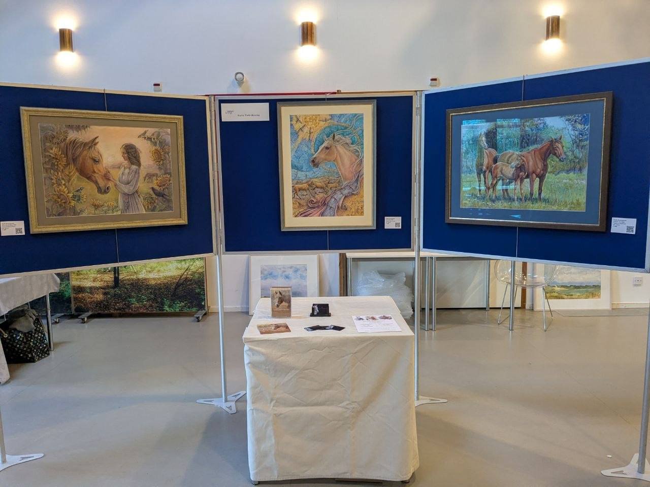 Art gallery exhibit featuring three framed horse paintings on blue display boards, with a table in front displaying photographs and informational papers.