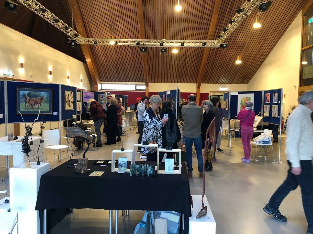 An art gallery or exhibition space with various artworks displayed on blue panels and tables. People are viewing and discussing art pieces, including paintings and sculptures, in a well-lit room with a wooden ceiling.