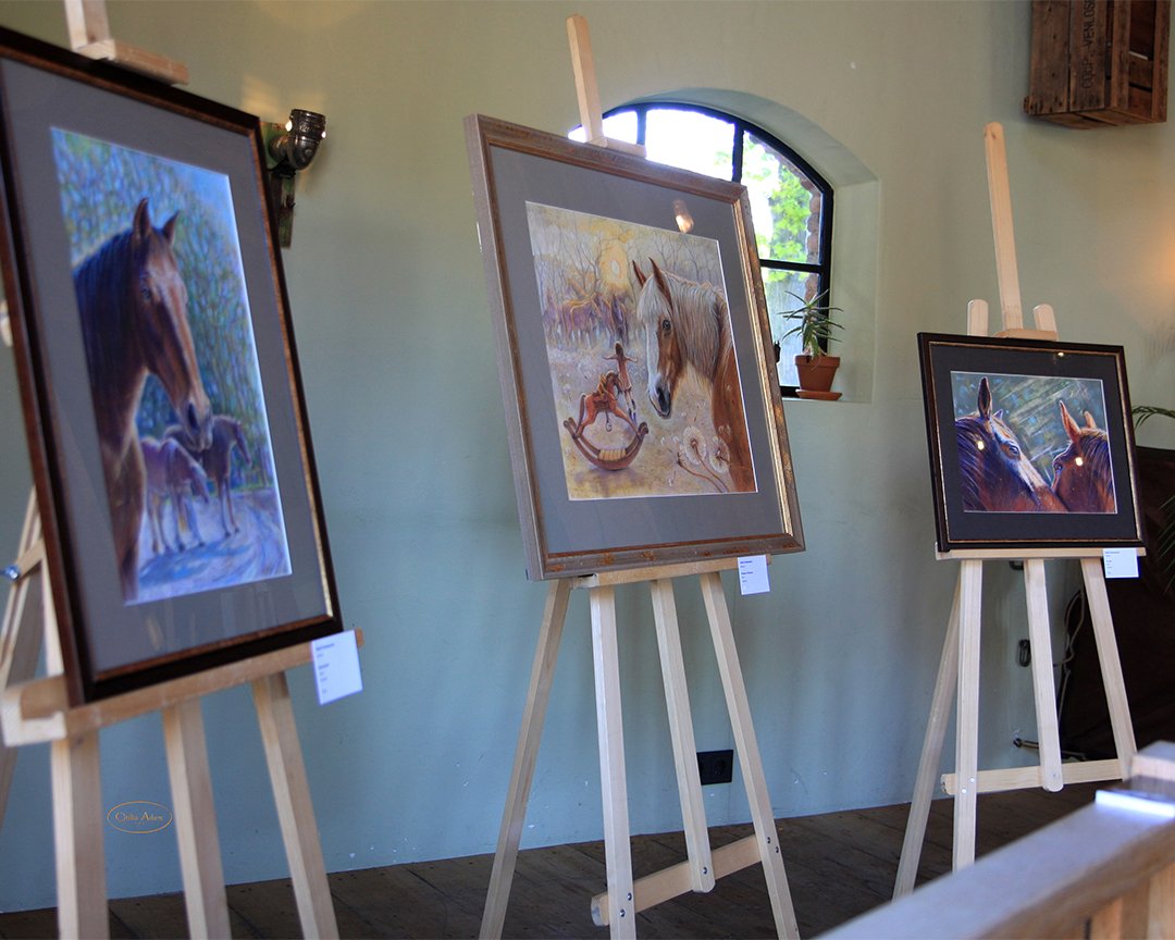 An art gallery displaying three framed, horse-themed paintings on wooden easels in front of a green wall with a small window. The paintings depict realistic horses, some with farm scenes, and are illuminated by gallery lighting.