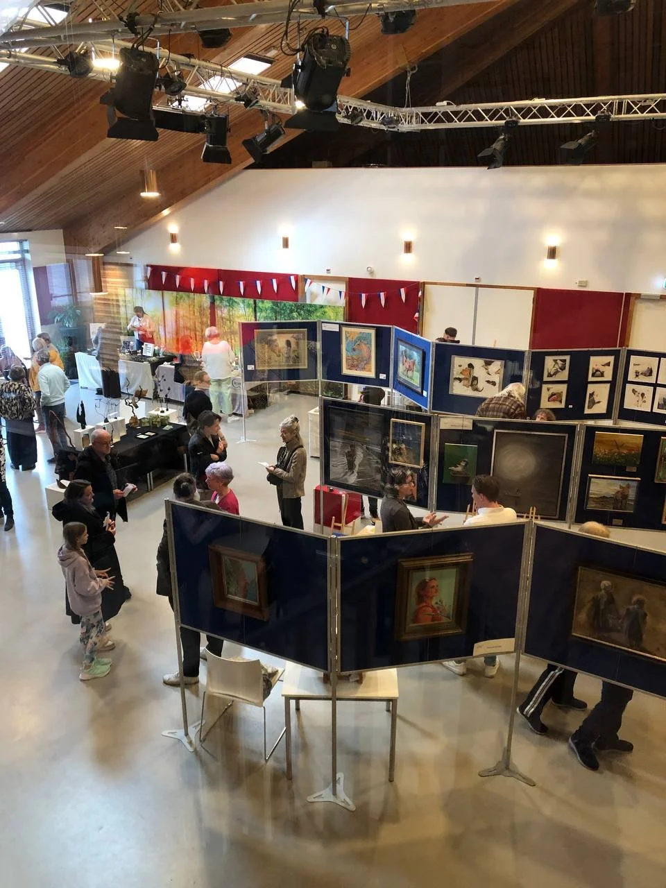 An indoor art gallery and auction event with people viewing paintings and artworks displayed on foldable partitions, a DJ or auctioneer at a table, and art pieces on display tables. The ceiling has wooden panels and stage lighting equipment.