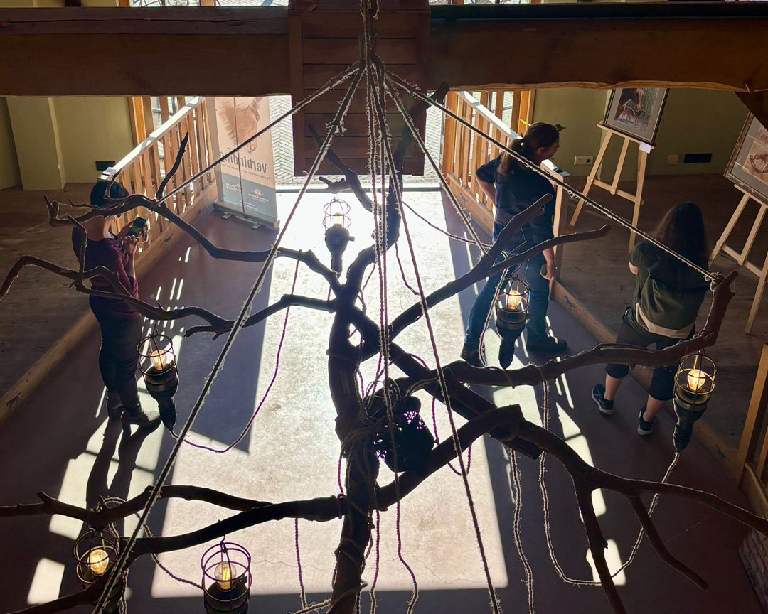 Interior of a building with a leafless tree sculpture hanging from the ceiling, surrounded by people looking at artwork displayed on easels.