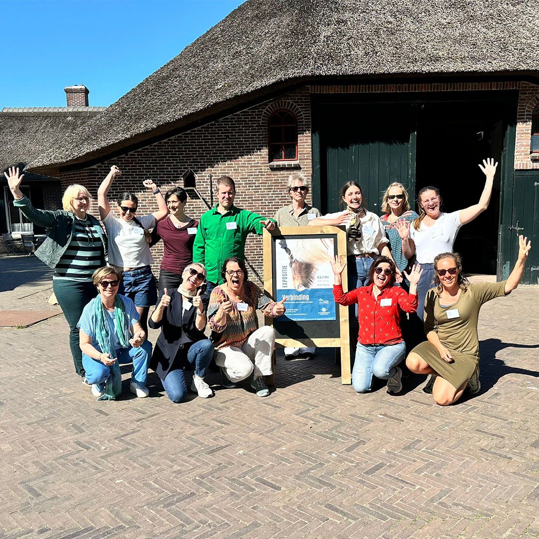 Group of people smiling and posing outdoors in front of a brick building, some giving thumbs-up and others raising their hands, during a sunny day.