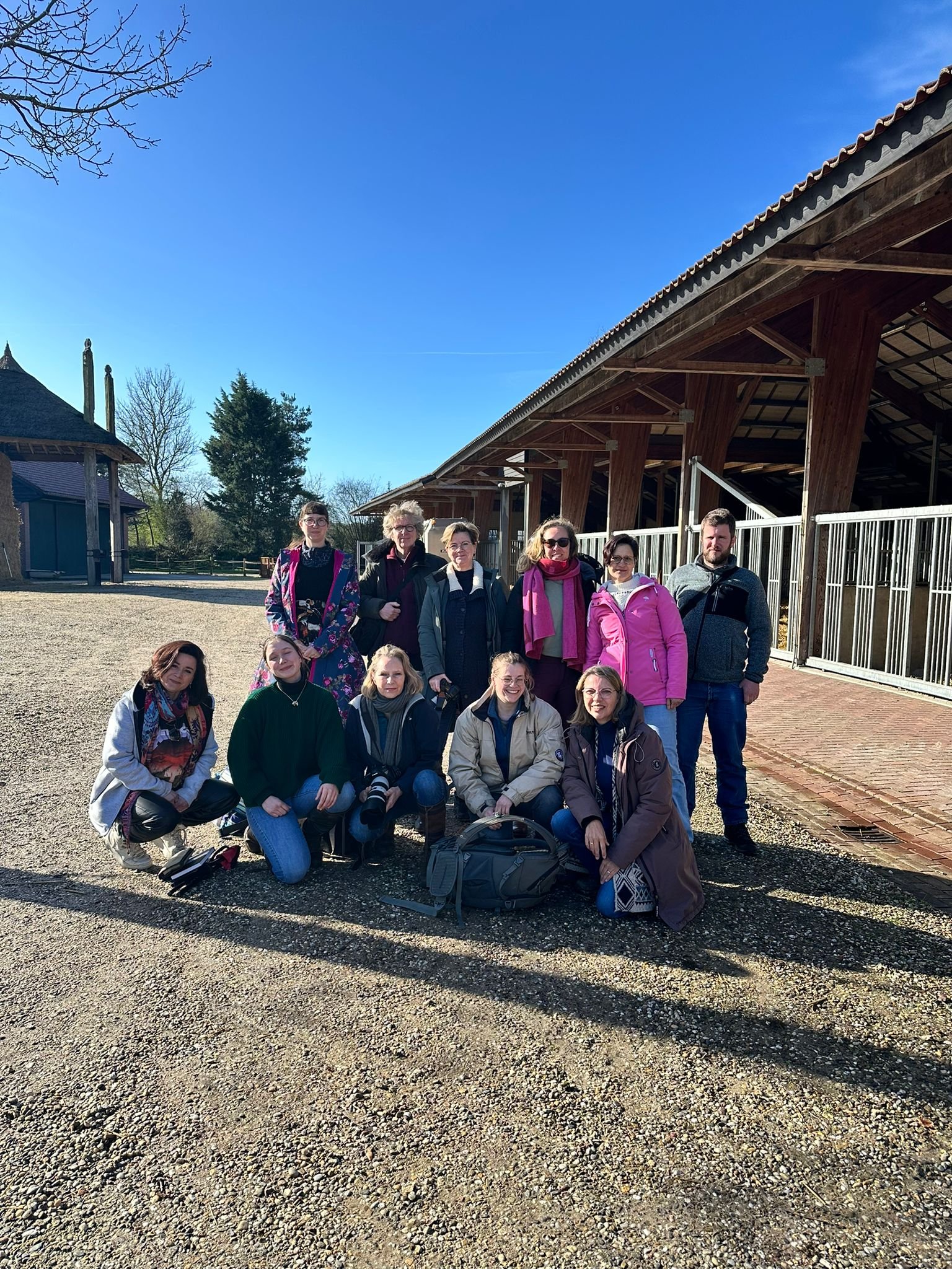 Group of people posing outdoors near wooden building on clear day with blue sky.