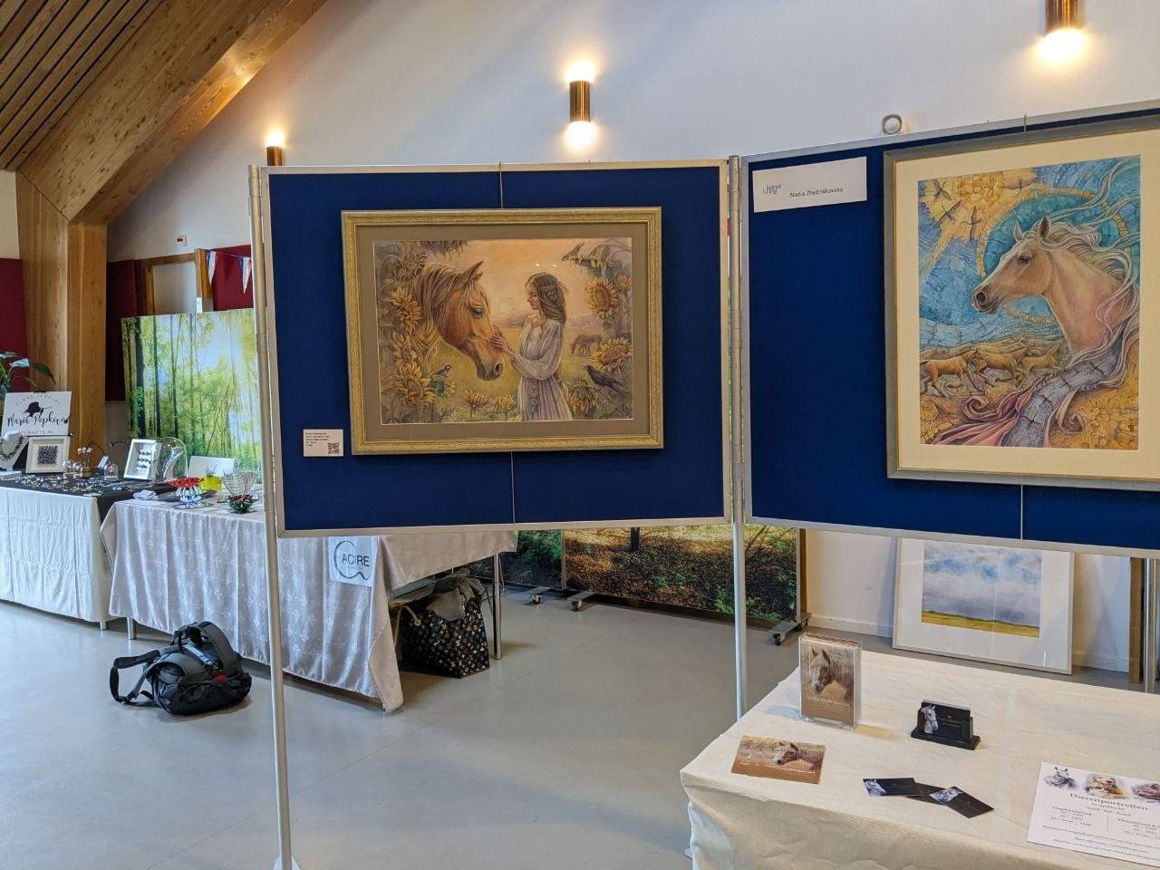 Art exhibition with two framed paintings of horses; the left painting depicts a woman and a horse in a landscape, and the right painting shows a horse with a divine background; tables with jewelry and brochures are in the background.