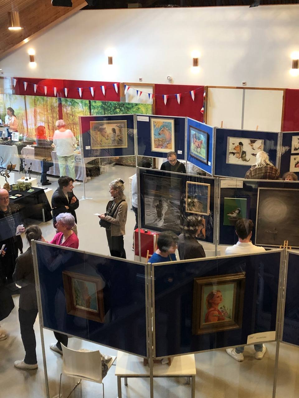 Art gallery with people viewing paintings and artwork on display panels, decorated with red, white, and blue bunting.