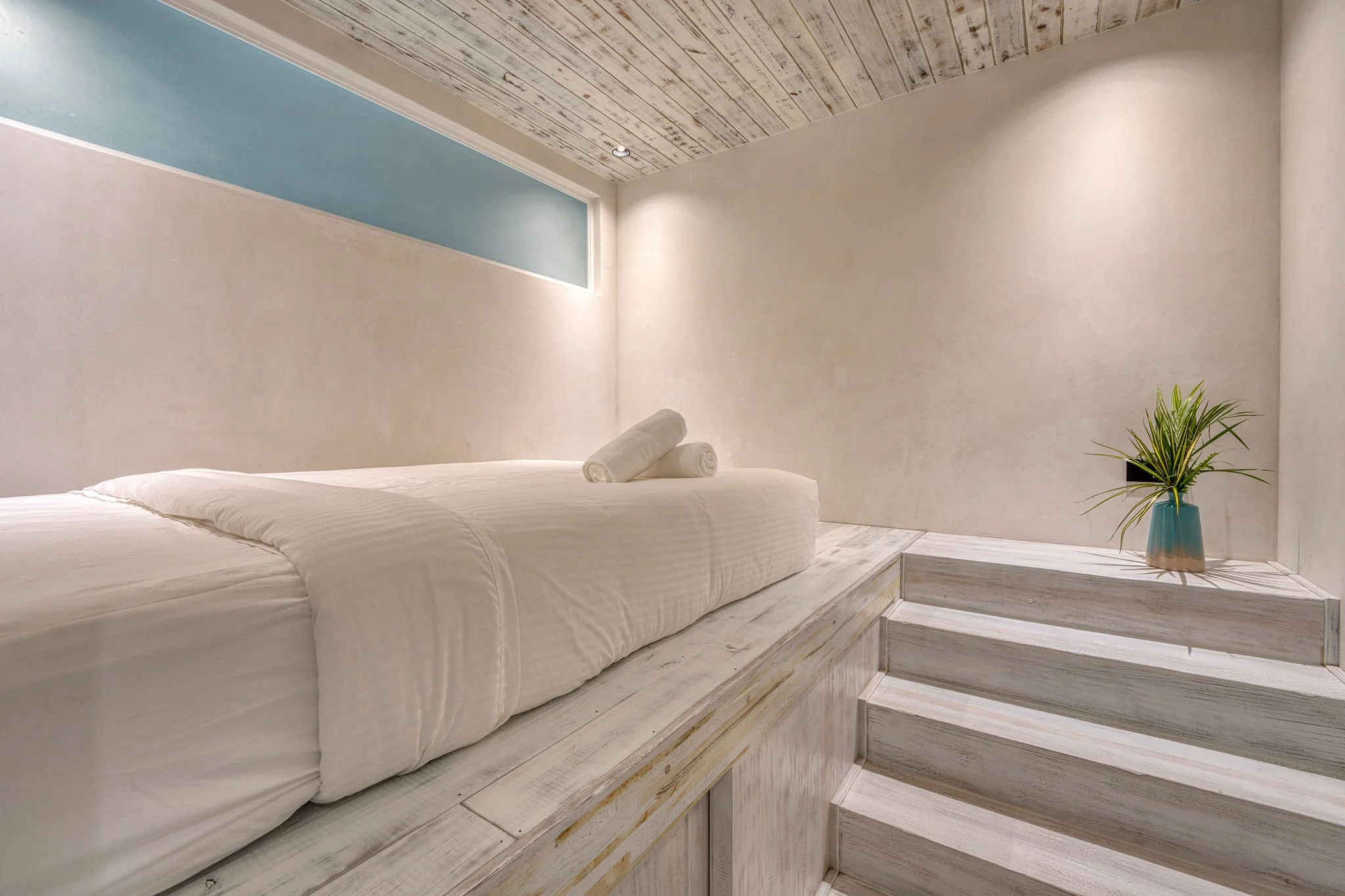 Minimalist bedroom with a platform bed dressed in white linens, a rolled towel, a small stair step, and a green plant in a blue vase. The room has white walls, a wood-paneled ceiling, and a narrow horizontal window.