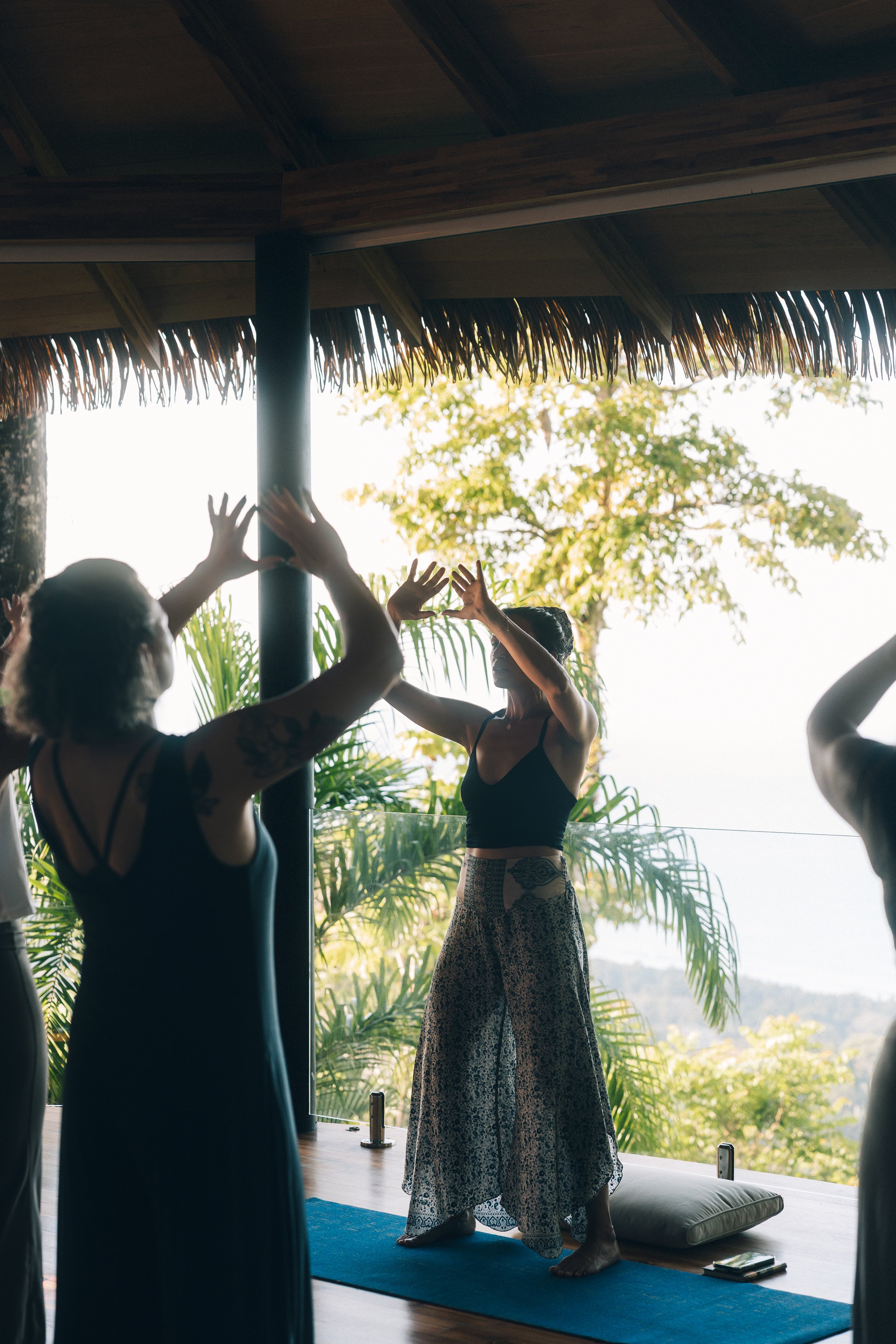 Group of women participating in a yoga session on a blue mat on a balcony with lush green trees in the background. The women are standing and reaching up with their arms, practicing yoga poses.