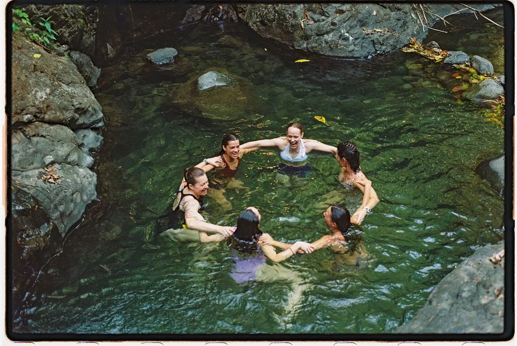 Six women holding hands and smiling in a natural pool surrounded by rocks and greenery.