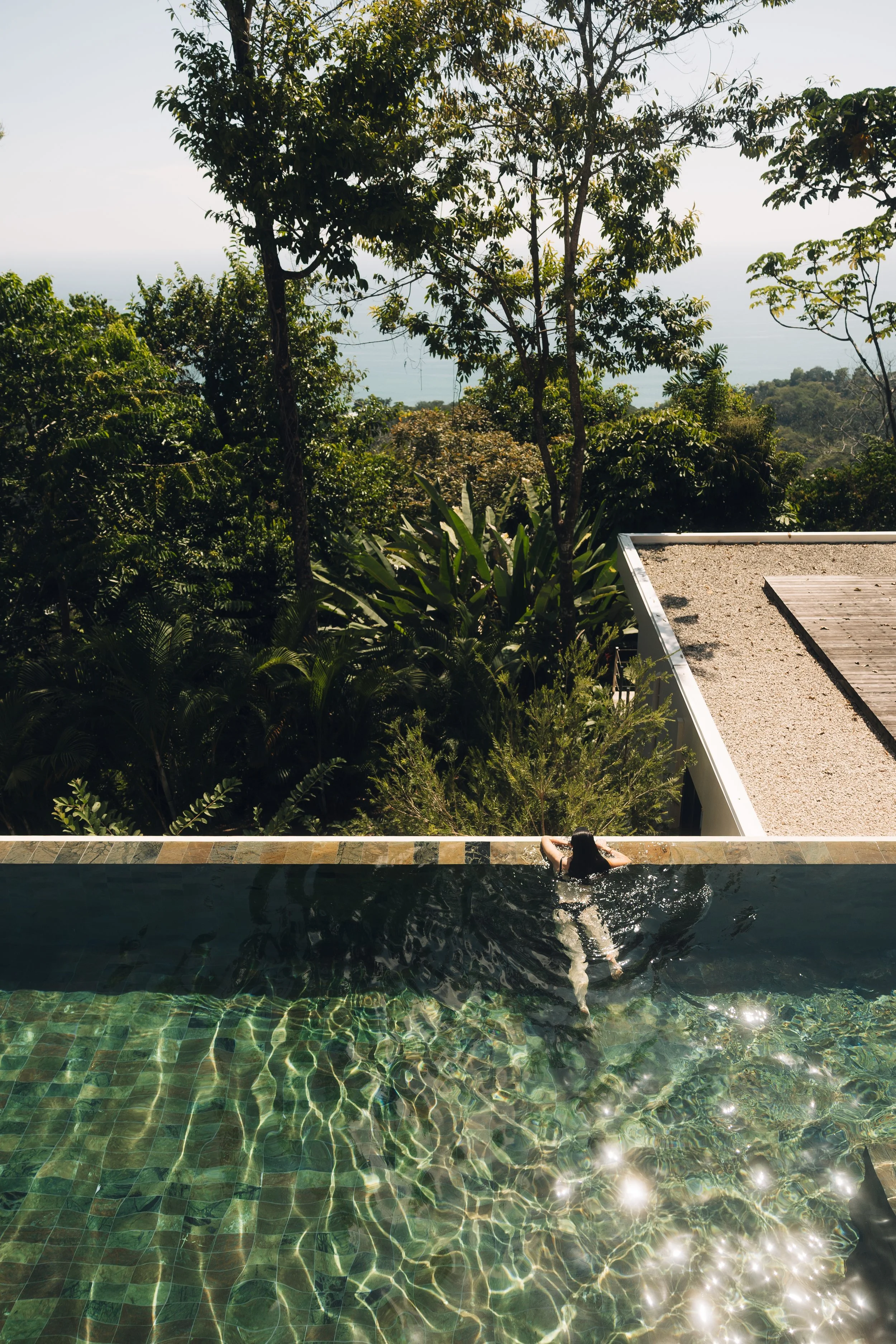 A woman relaxing in a swimming pool with lush green trees and a distant view of hills or mountains in the background.