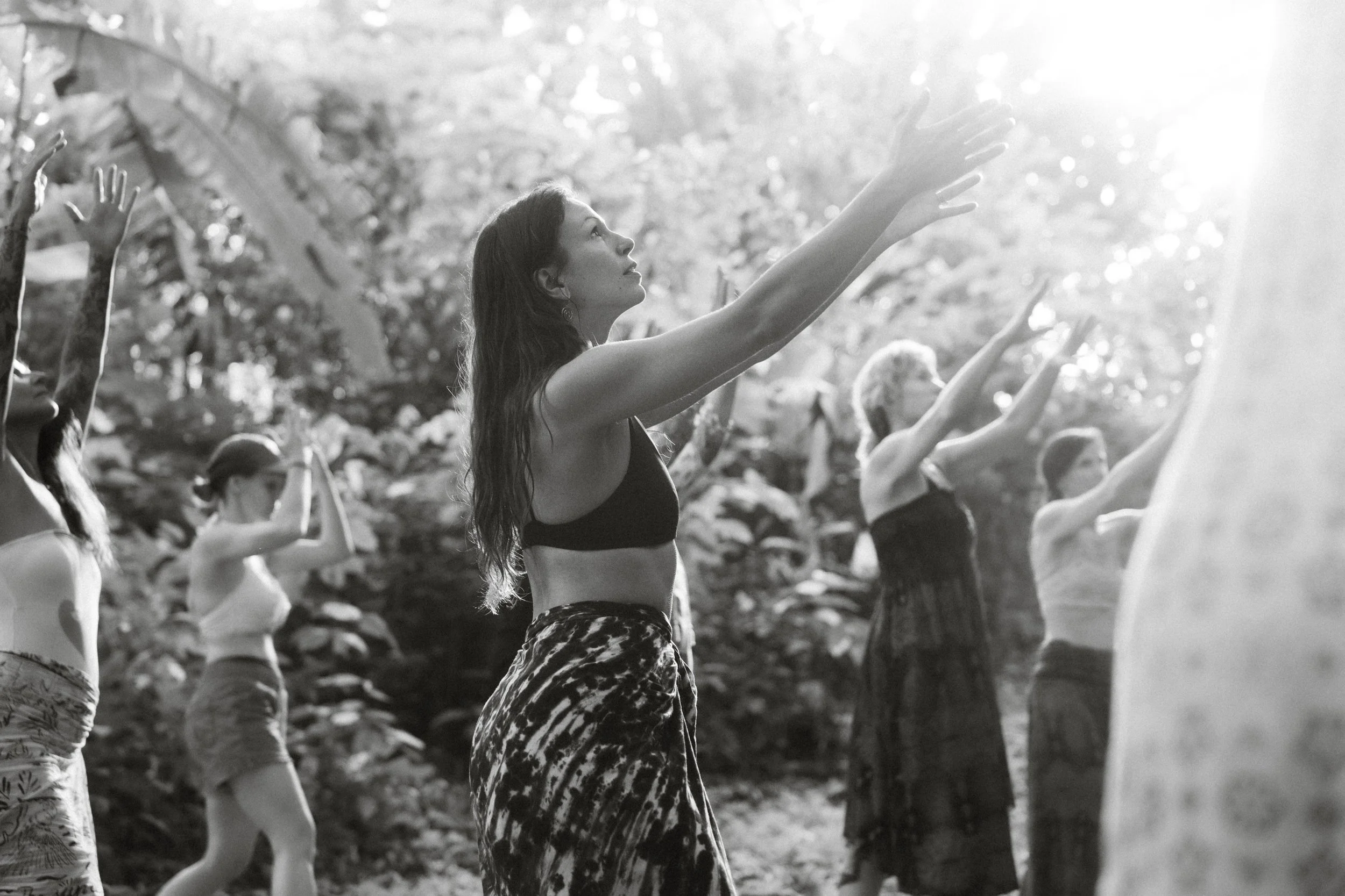 A group of people practicing yoga outdoors in a forest, reaching up with their hands during a yoga session.