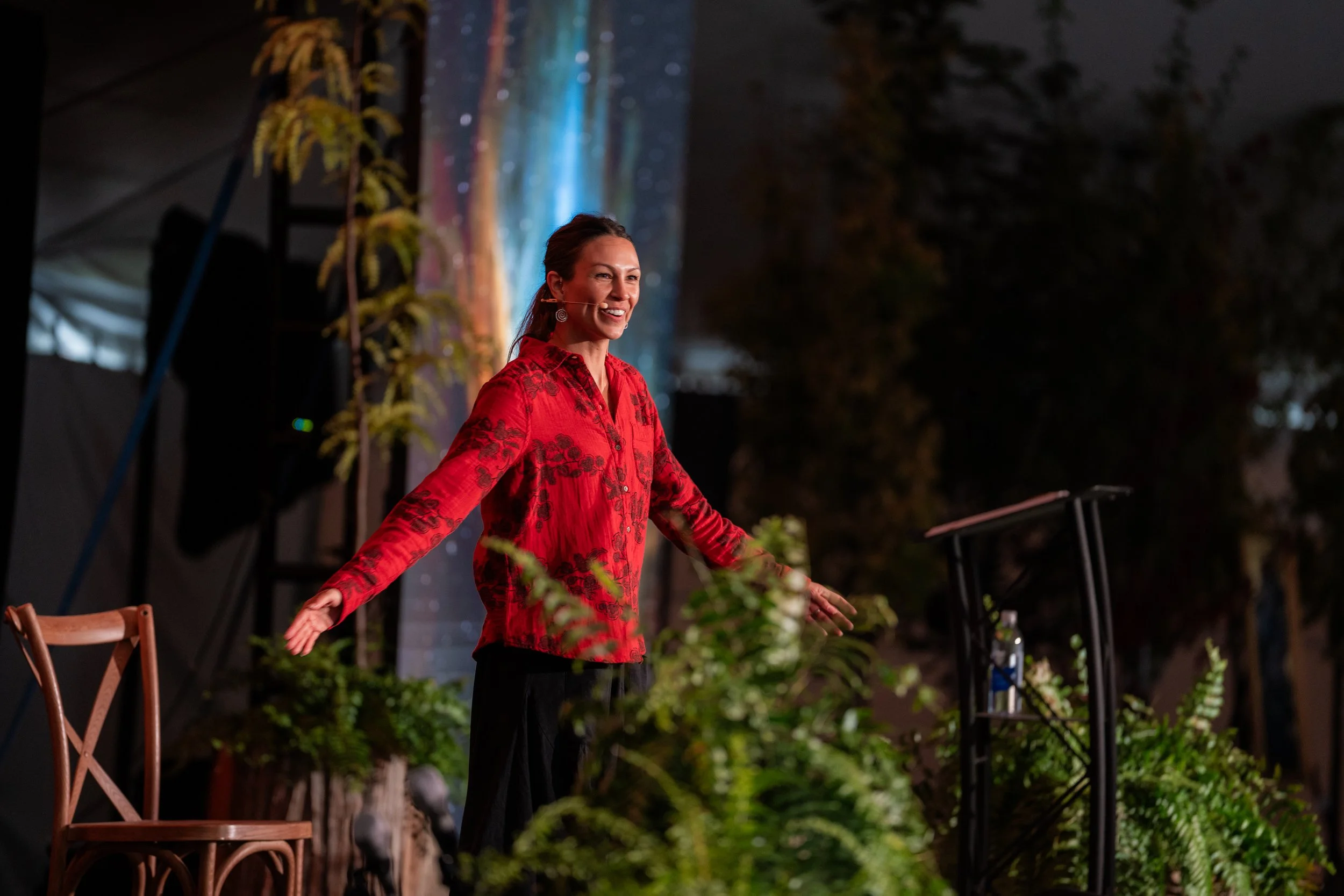 Stephanie Nosco standing, smiling, wearing a red patterned blouse, with her arms outstretched teaching at New Human Event
