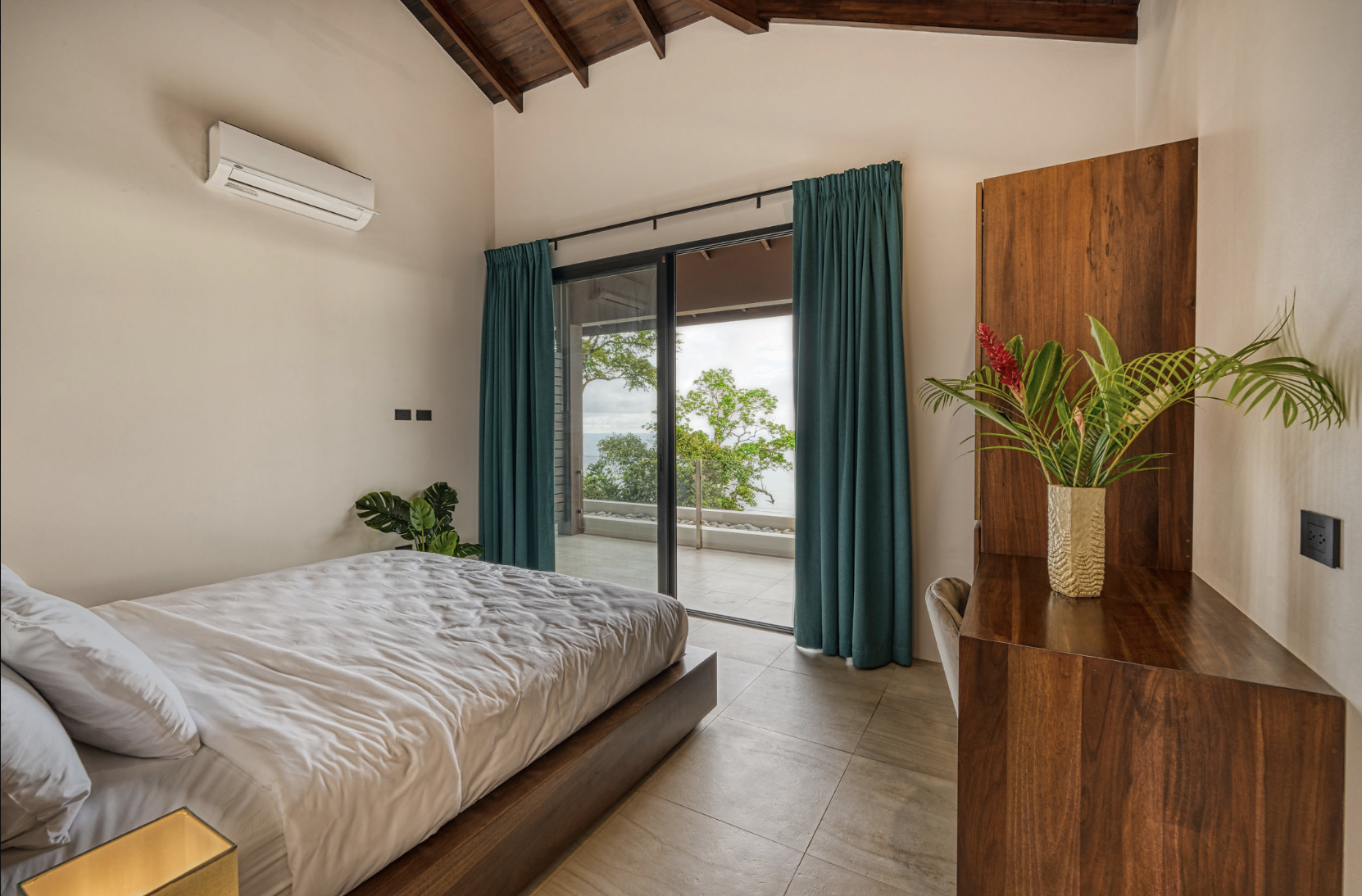 A bedroom with a wooden bed frame and white bedding, a potted plant with large green leaves on the nightstand, green curtains hanging in front of a glass sliding door, outdoor trees visible through the glass, and a wooden dresser with a tray and plan