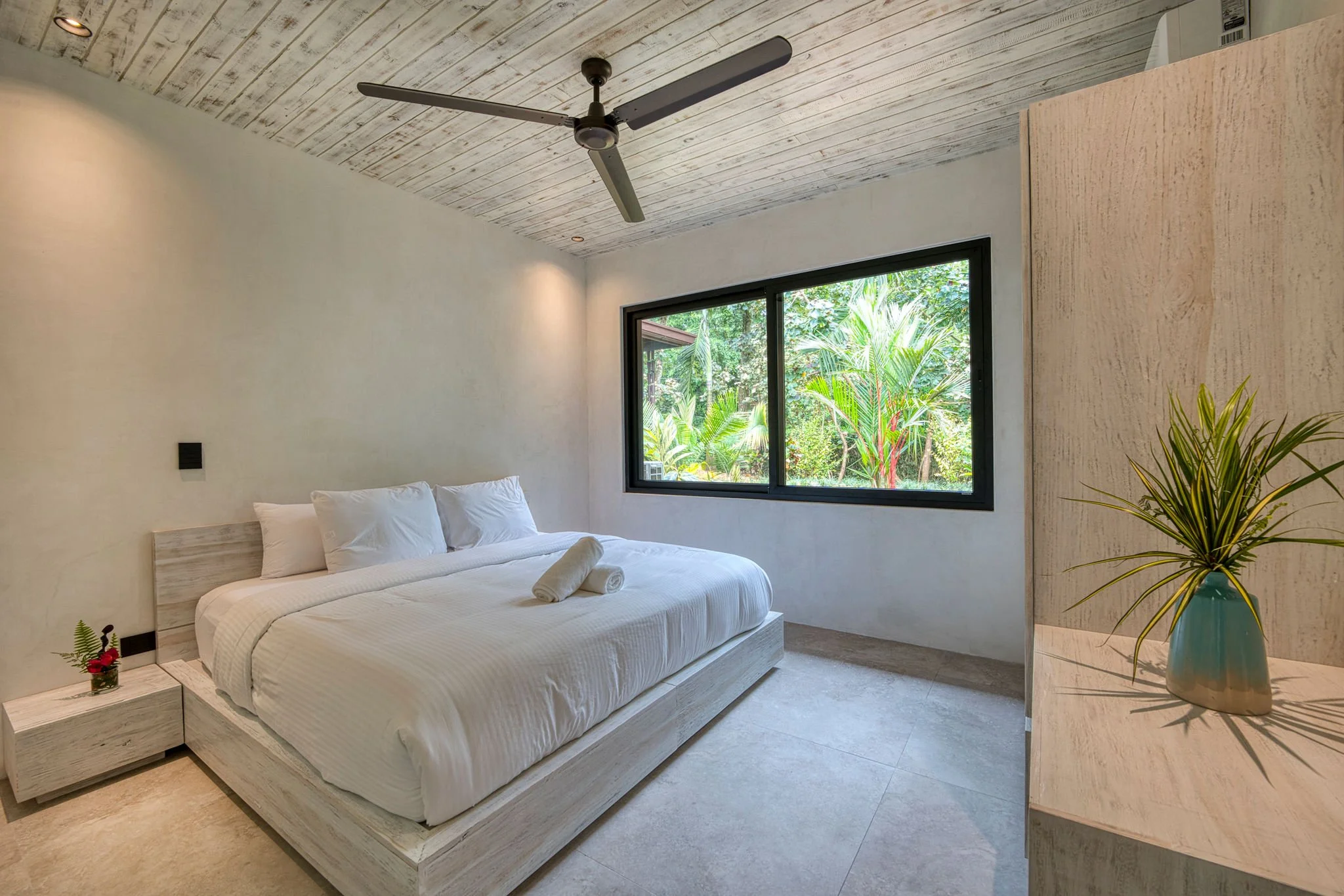 A bedroom with a white bed, pillows, and a rolled towel. There is a small nightstand with a flower vase. A large window shows green trees outside, and the ceiling has a fan and recessed lighting.