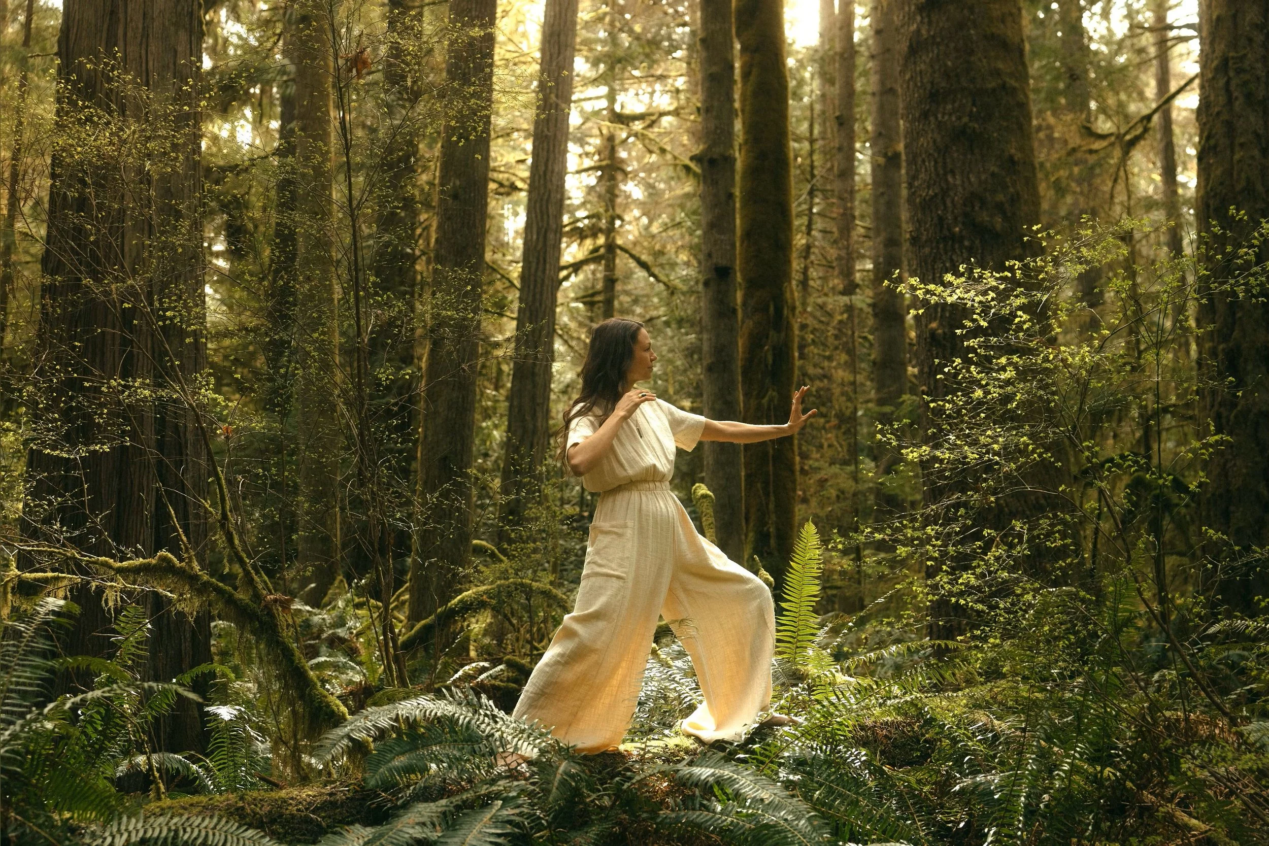 Stephanie Nosco standing in a qi gong form in the forest. She is wearing wide legged pants, and the outfit is a wheat coloured linen one piece