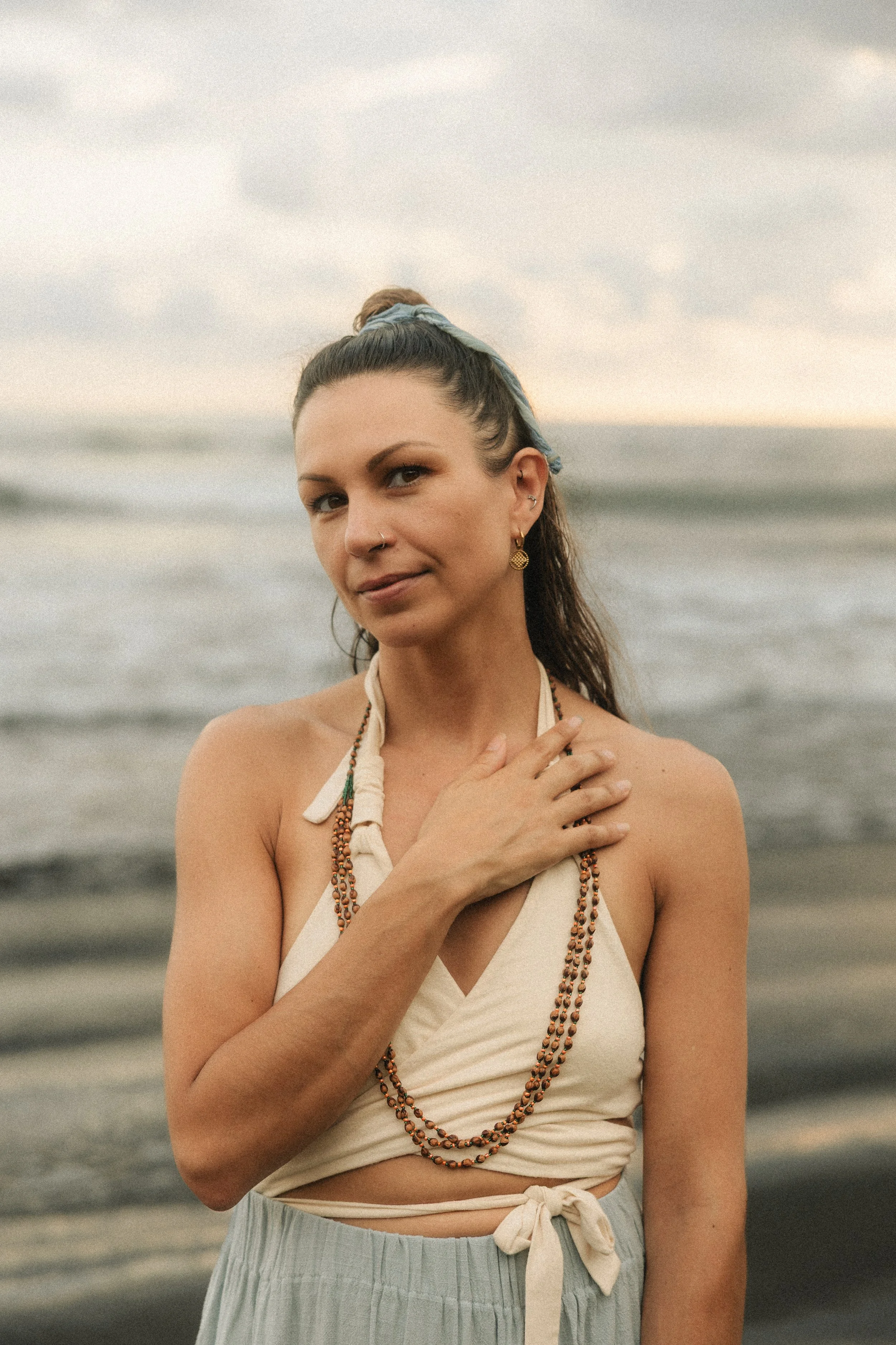 A woman with dark hair in a high bun, wearing a beige top, layered necklaces, and earrings, standing on a beach with ocean waves in the background.