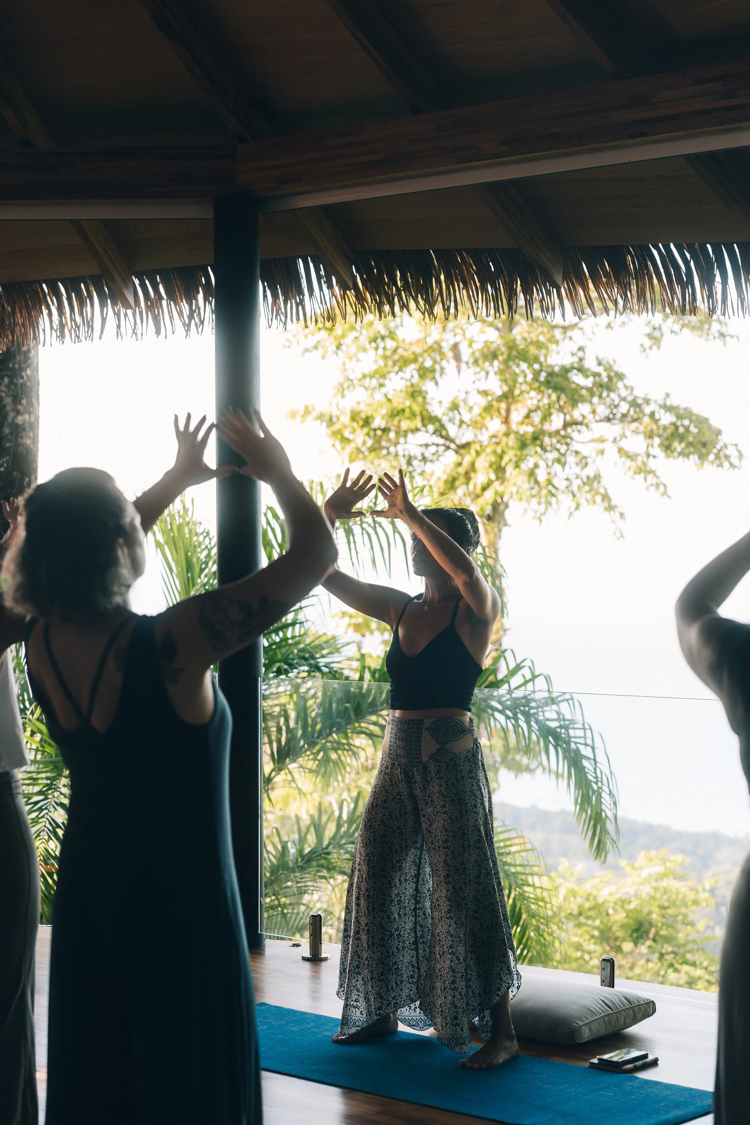 People practicing yoga on a blue mat in a thatched-roof space surrounded by lush tropical plants and trees, with a view of distant hills or mountains.