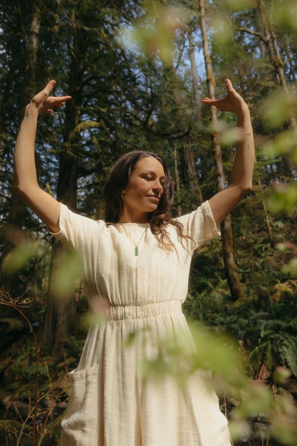 Stephanie Nosco has her hands raised overhead in a qigong form. She is surrounded by old growth forest. She is wearing a short sleeved wheat coloured linen one piece outfit.
